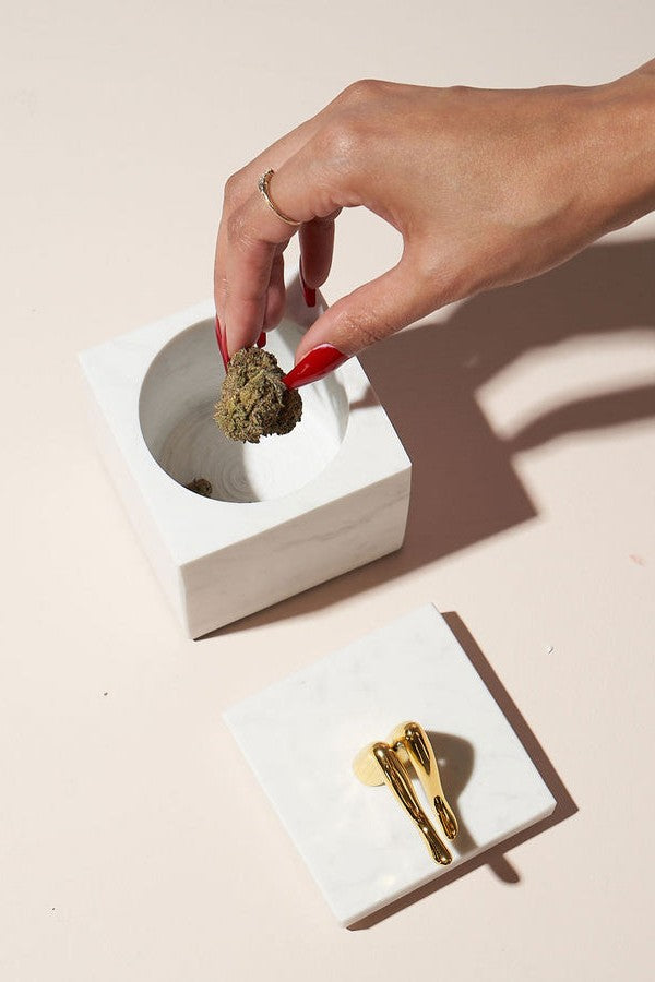 Hand placing cannabis bud into an open white container with rolling papers nearby, bathed in soft light.