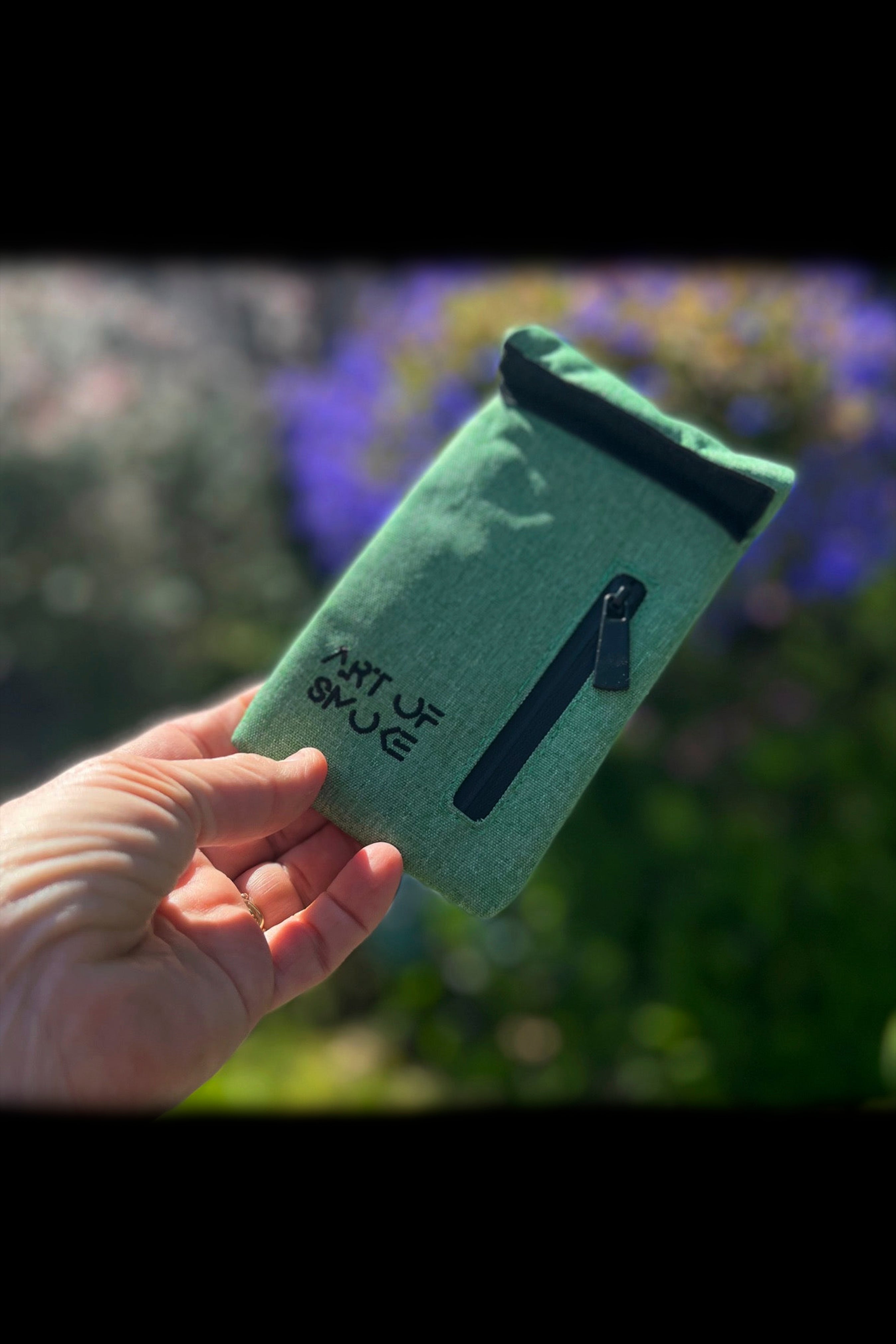 A hand holds a green, zippered smell-proof bag with “ART OF SMOKE” embroidered on it, set against a blurred, natural outdoor background.