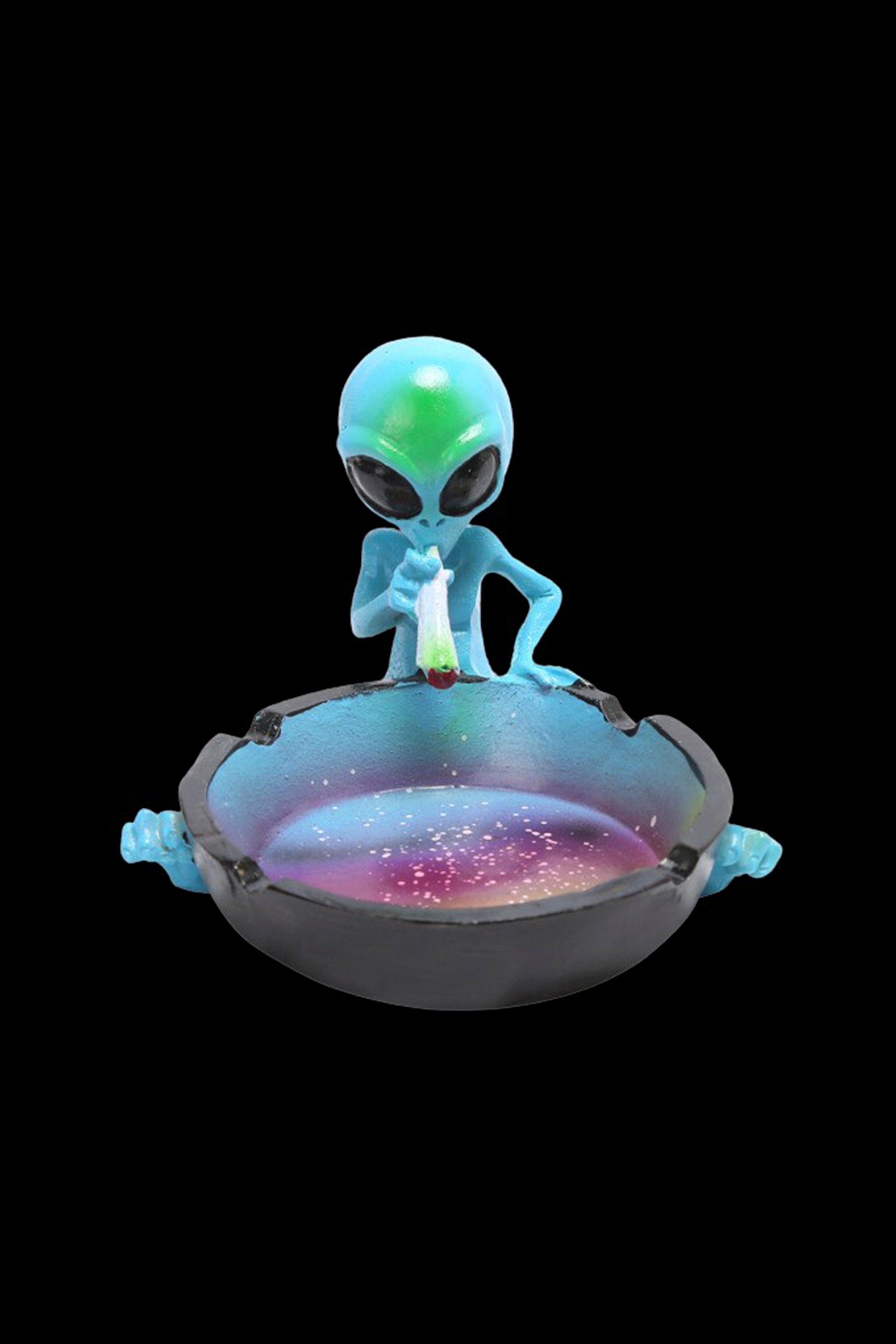 A vibrant, iridescent glass ashtray sculpted as an alien spaceship, topped with a miniature, turquoise alien figurine appearing to smoke cannabis.