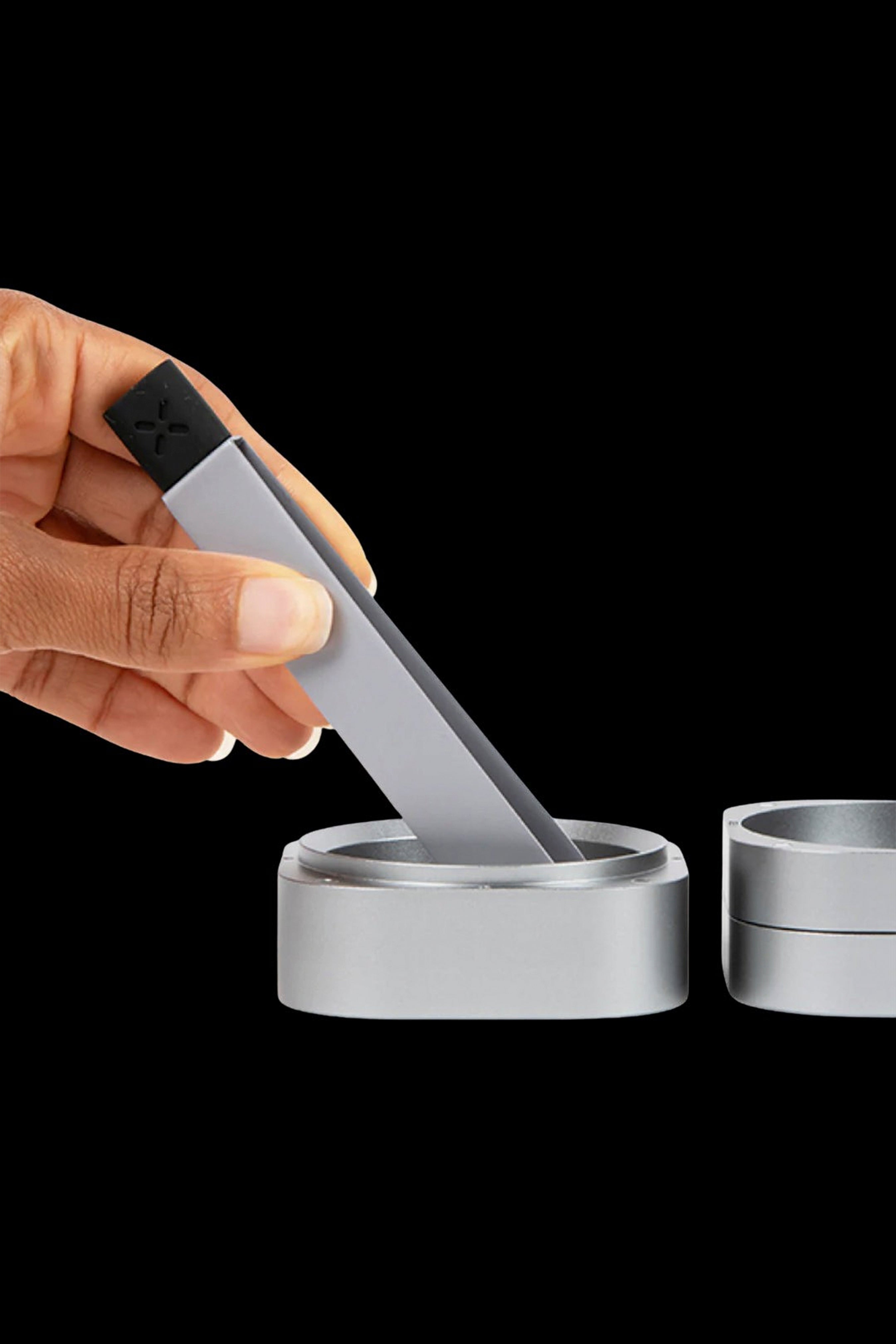 Hand using a metal tool to collect cannabis concentrate from a metallic container, with a stack of similar containers blurred in the background.