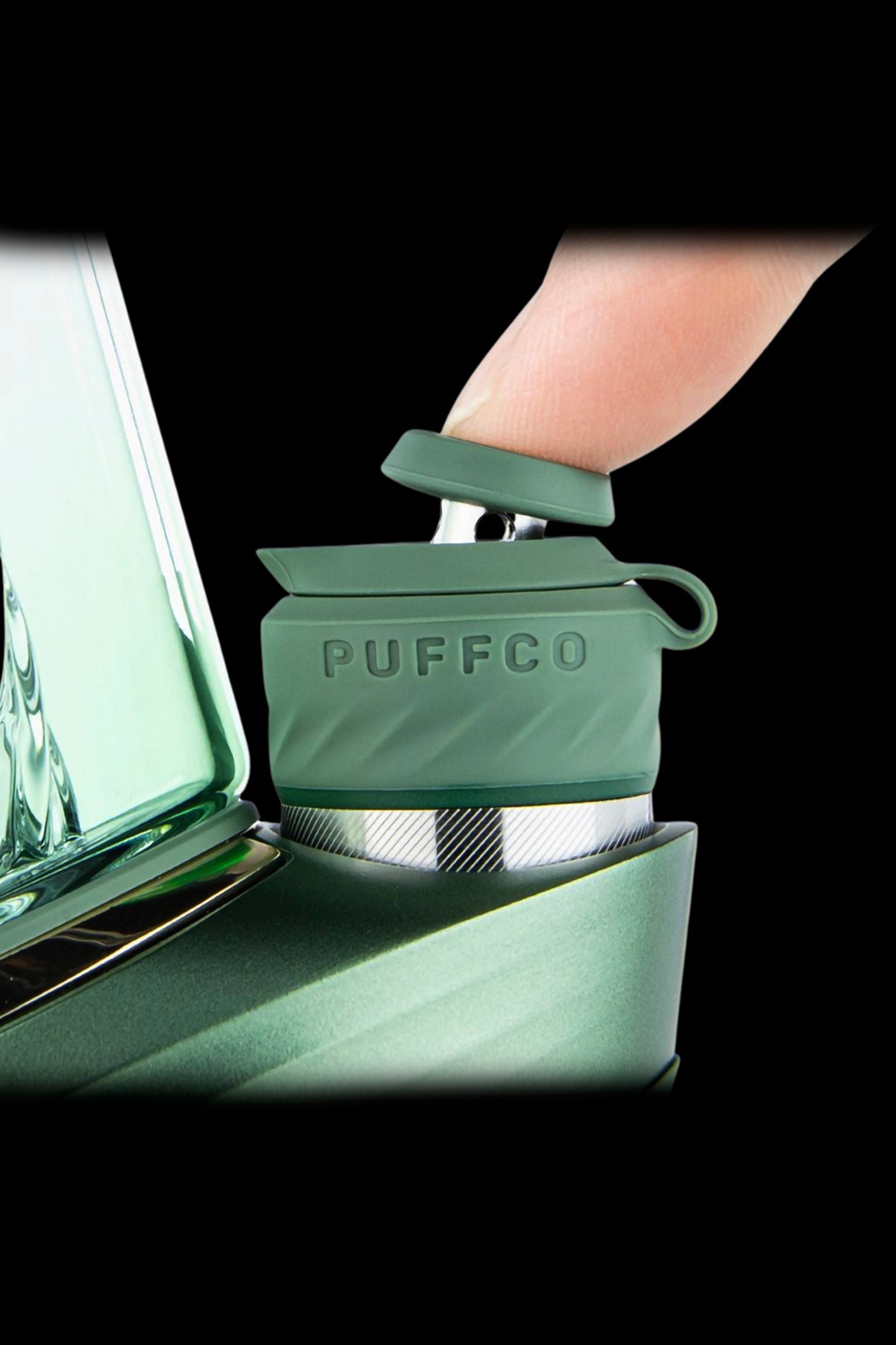 Close-up of a green Puffco Peak atomizer with a hand gently touching it, against a blurred green background.