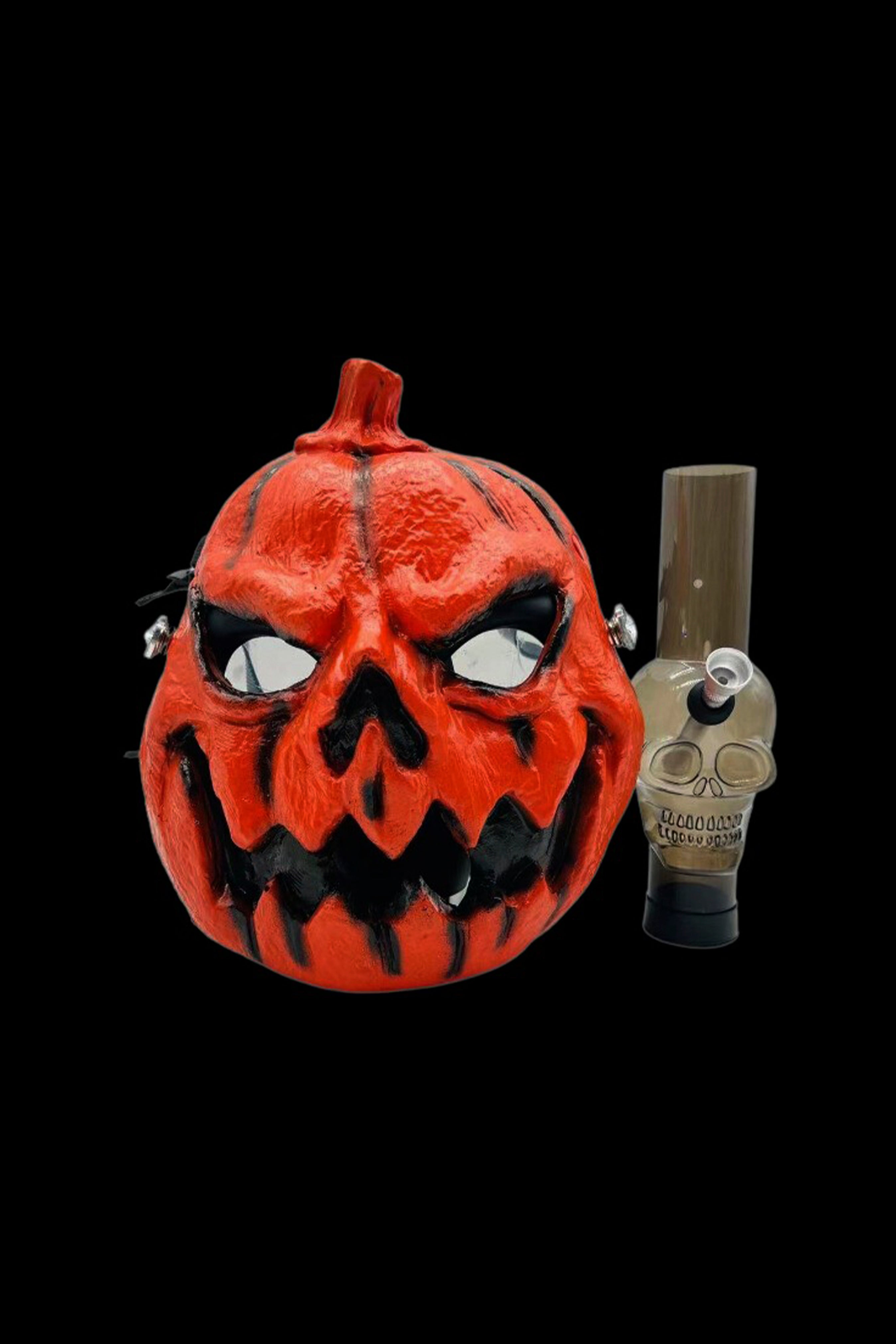 A vibrant orange jack-o'-lantern mask positioned beside a glass cannabis oil burner, both set against a black background.