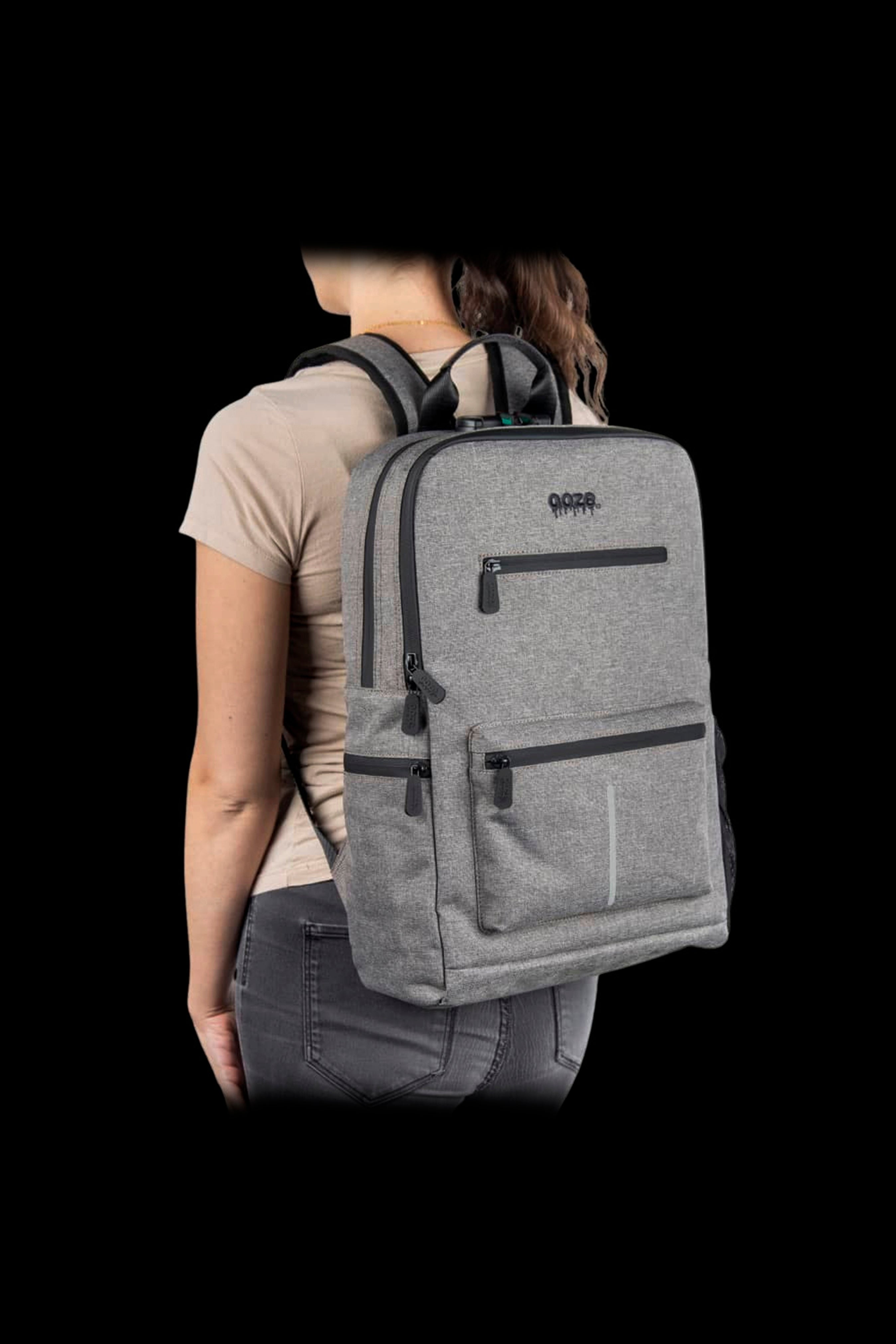 Rear three-quarter view of a woman wearing the Ooze Traveler Smell Proof Backpack in light gray against a black background.