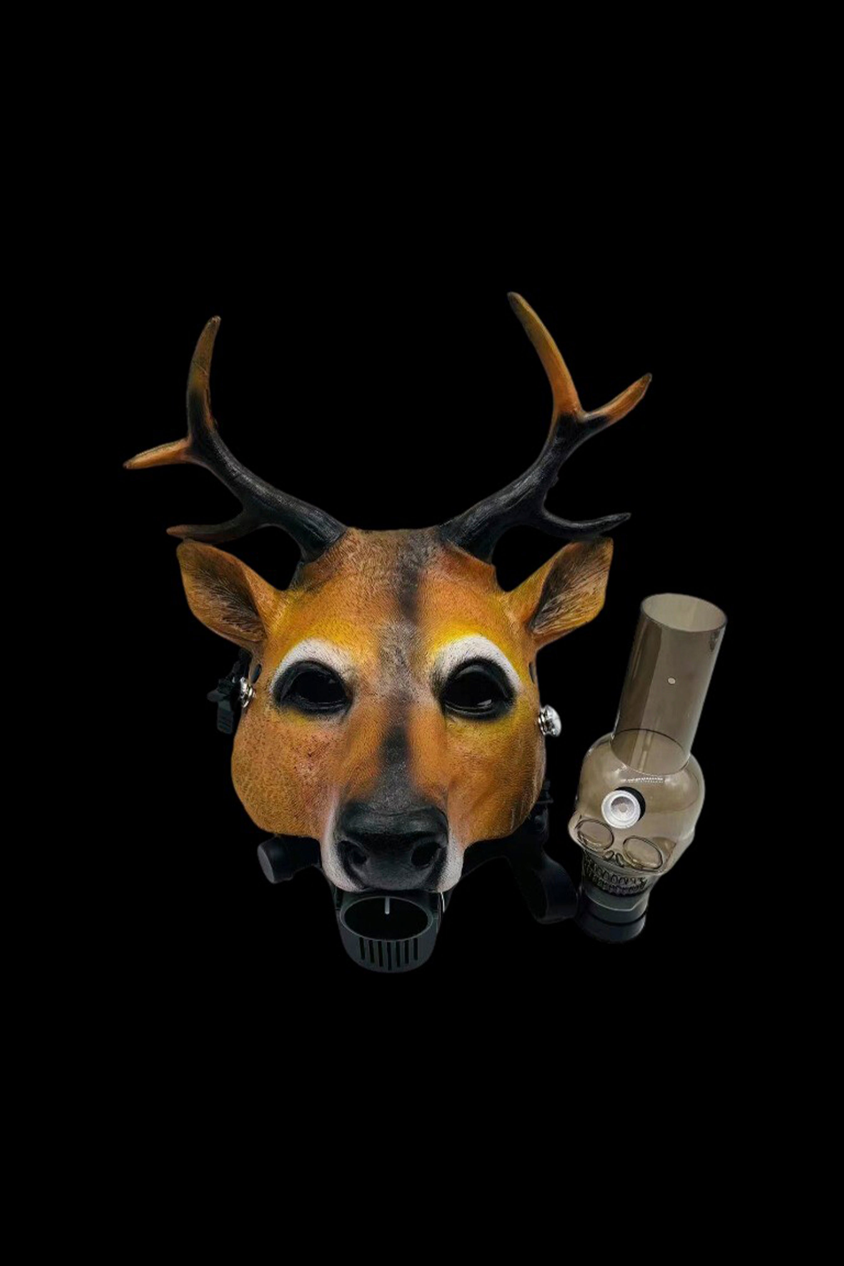 A deer mask with glowing eyes holds a glass cannabis oil rig in its mouth against a black background.
