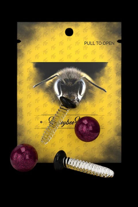 Close-up of a bee’s face over a patterned yellow background with cannabis accessories including a metal and glass smoking piece and red resin pellets, labeled “keybee”.