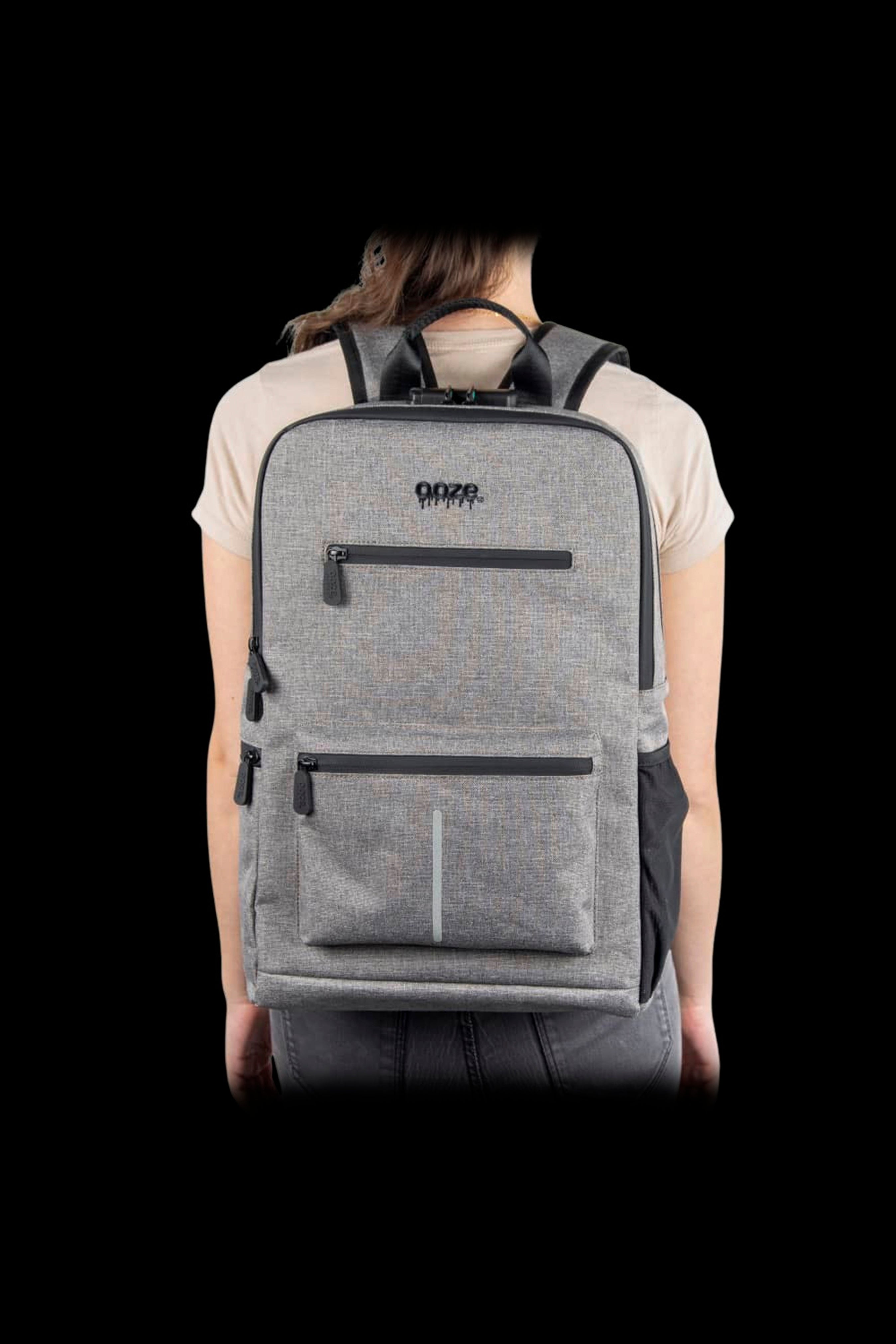Rear view of a person wearing the gray Ooze Traveler Smell Proof Backpack against a black background, showing padded straps and front zip pockets.