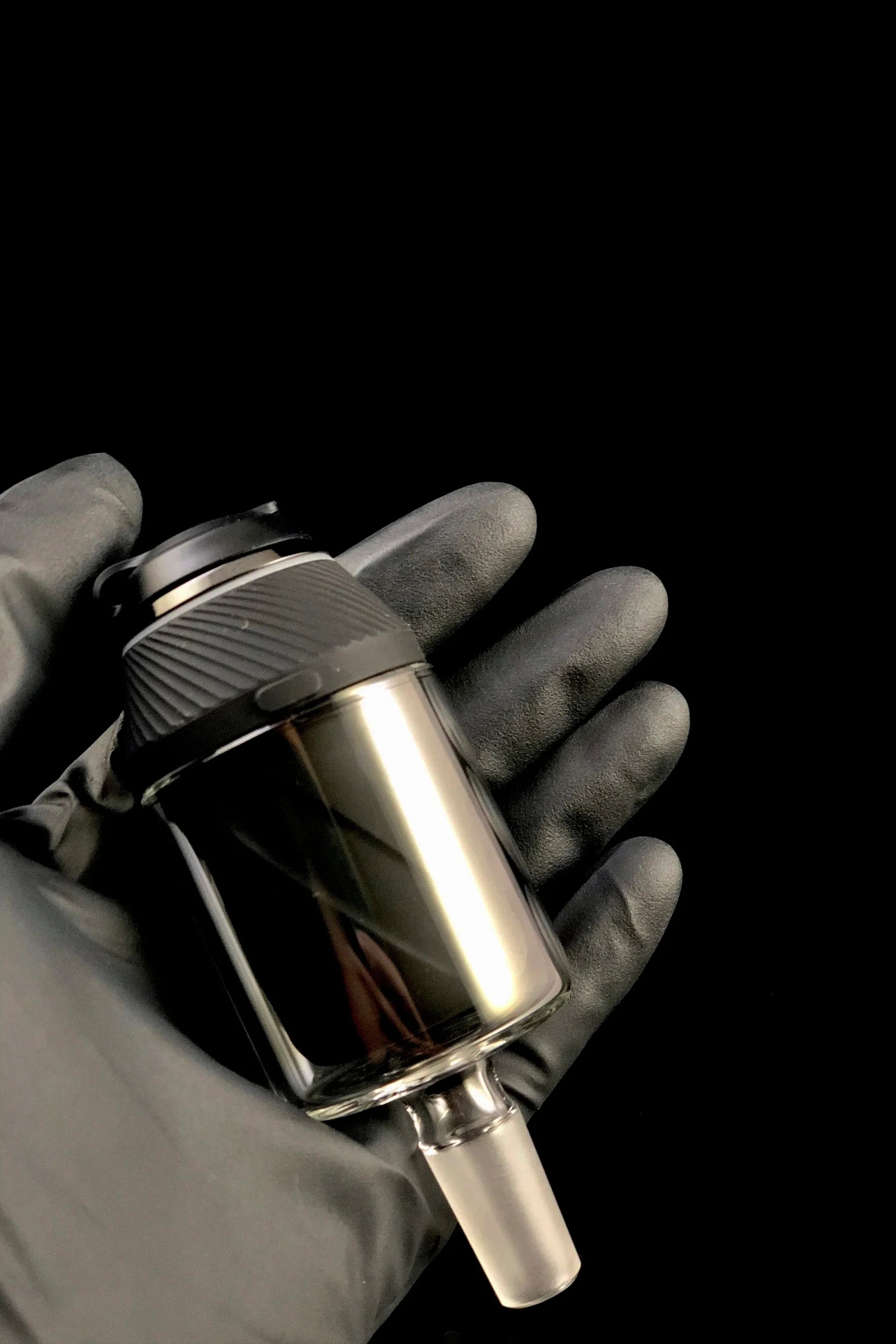 A gloved hand holds a sleek, metallic cannabis oil rig with a glass chamber.