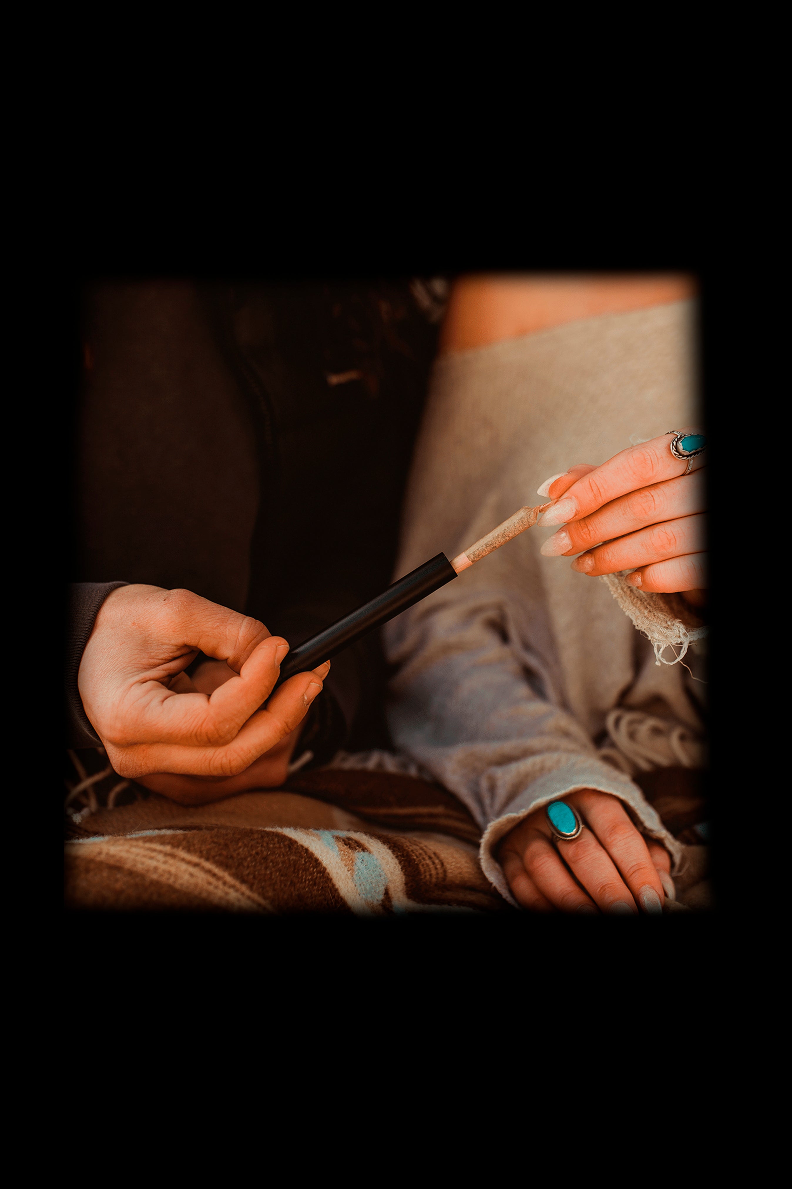 Close-up of hands preparing a cannabis joint with a metal packing tool, turquoise rings visible.