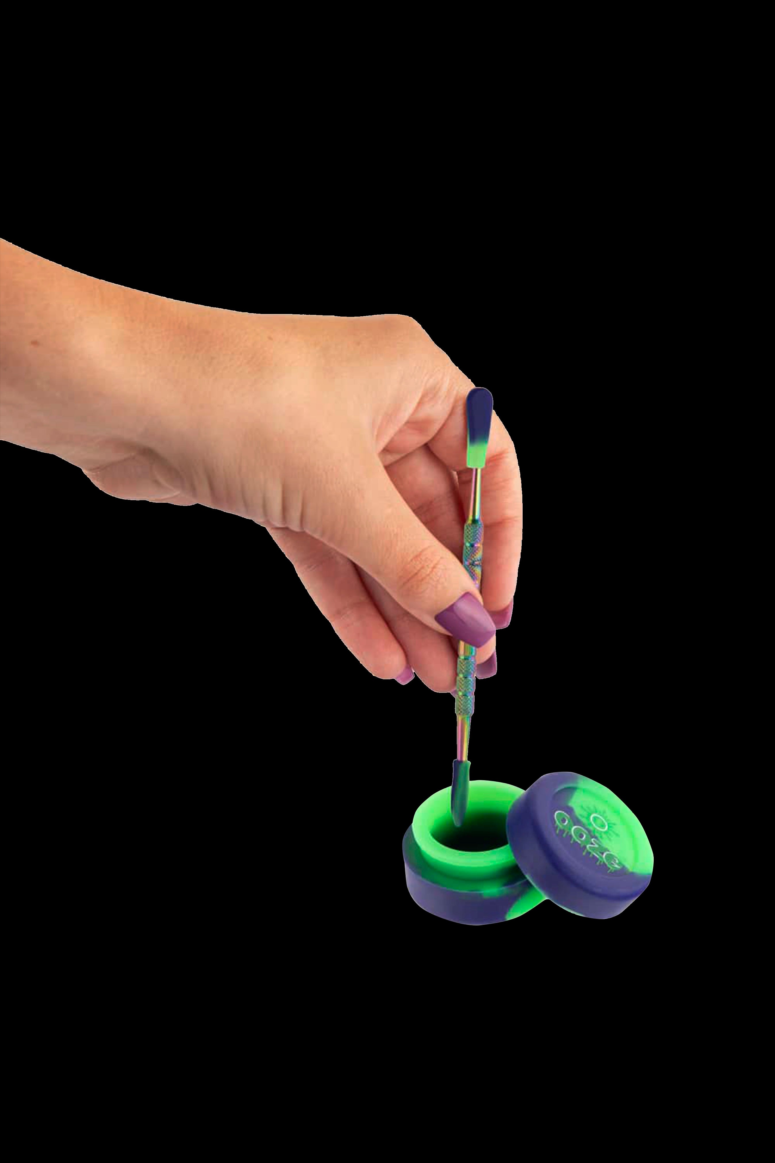A hand holding a dab tool over a silicone container, both with iridescent purple and green coloring.