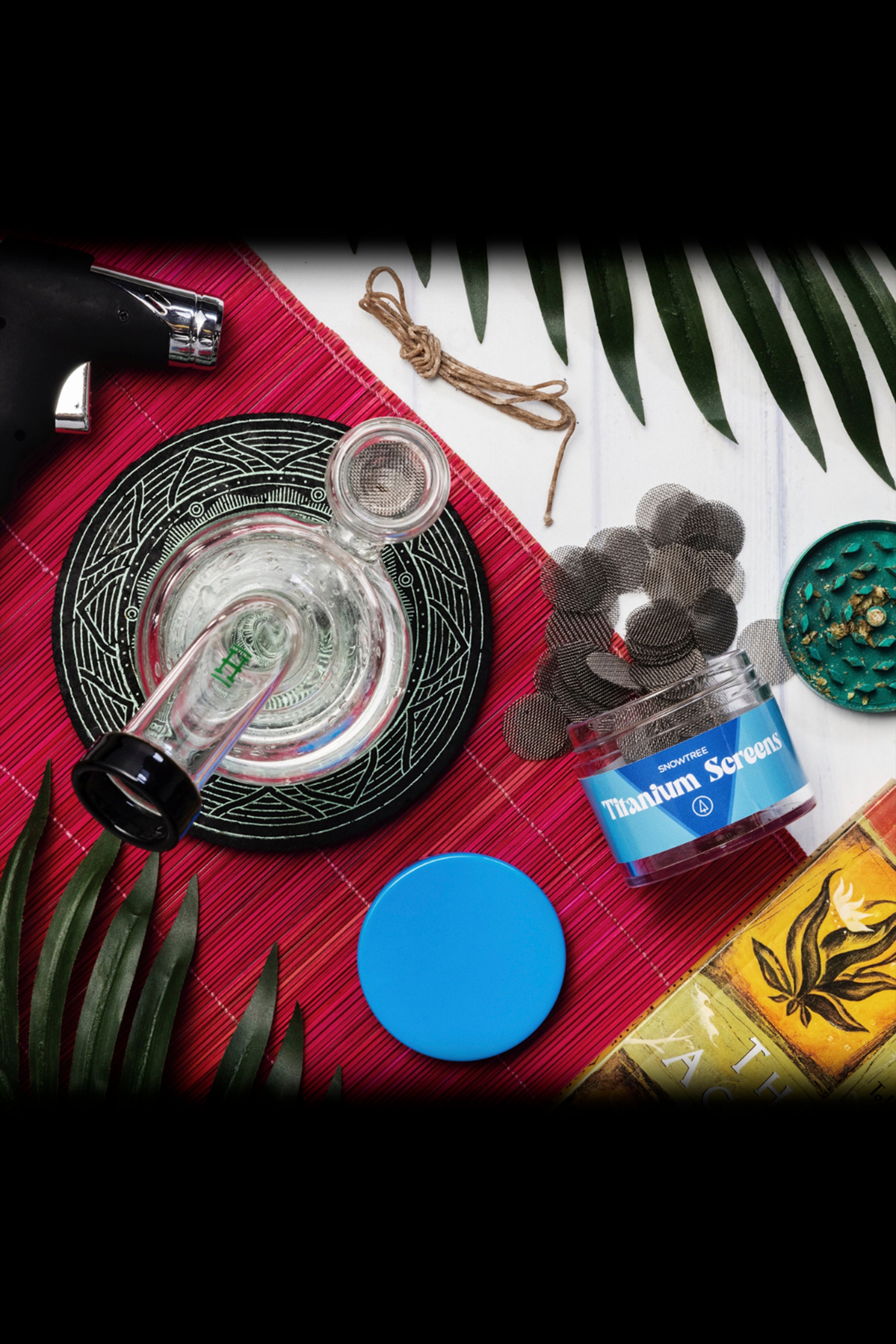 Overhead shot of cannabis accessories including a glass pipe, titanium screens, lighter, and rolling papers arranged on a patterned mat with tropical leaves.