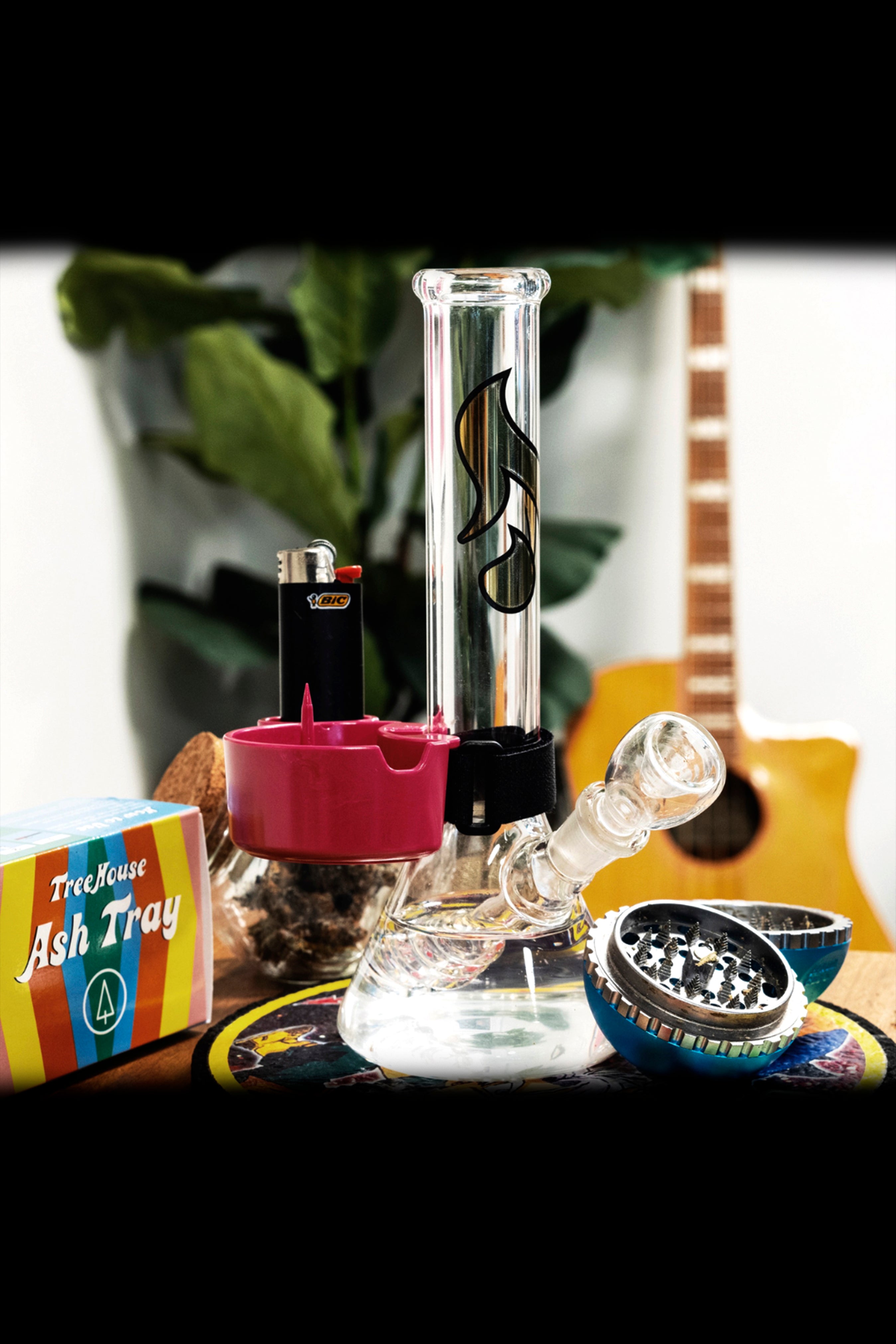 A glass bong with a pink accent piece, lighter, grinder, and ashtray on a patterned surface, with a blurred plant and guitar in the background.