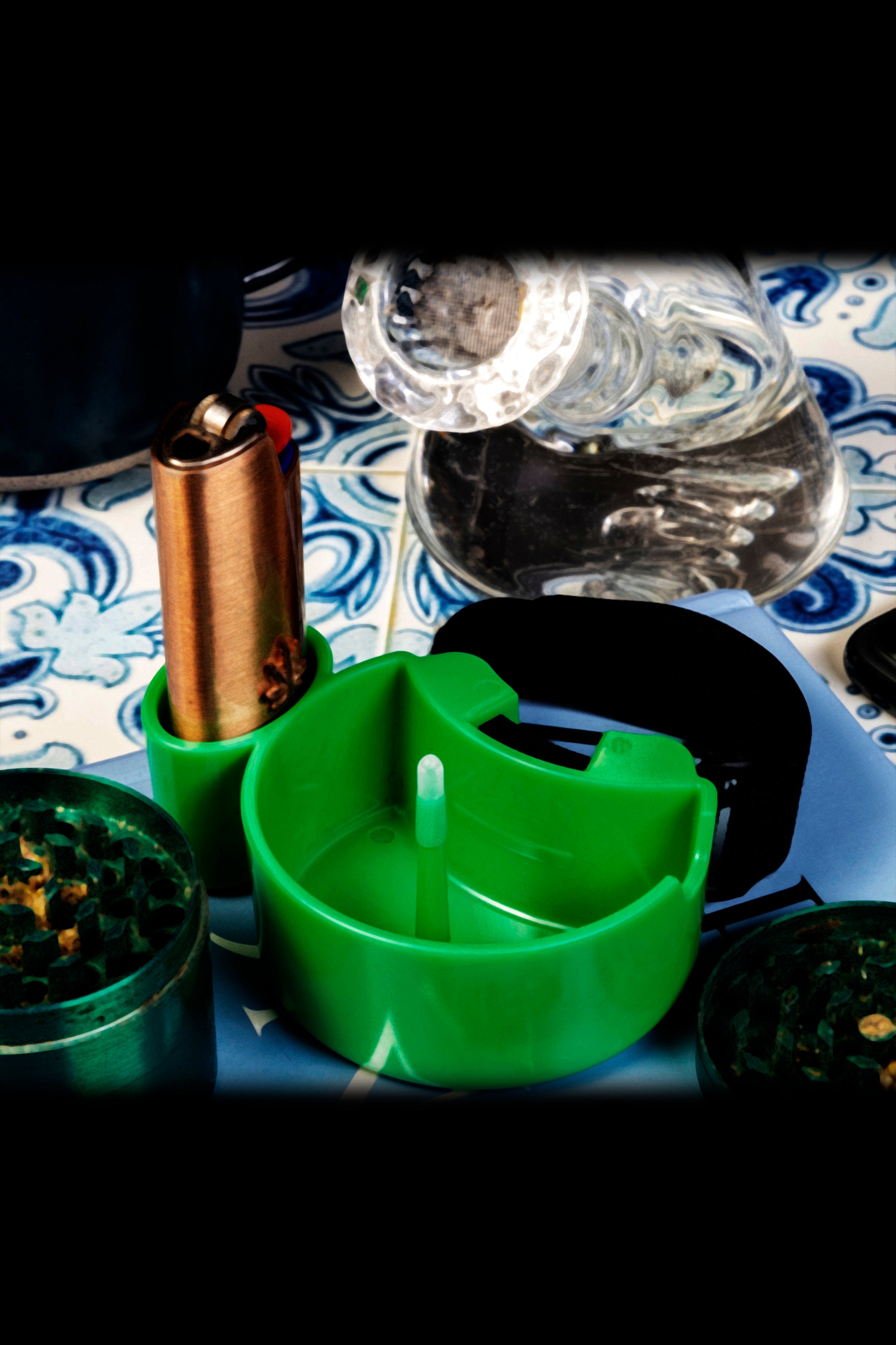 Cannabis accessories including a green grinder, small containers of bud, a lighter, and water bottle arranged on a patterned surface with dramatic lighting.