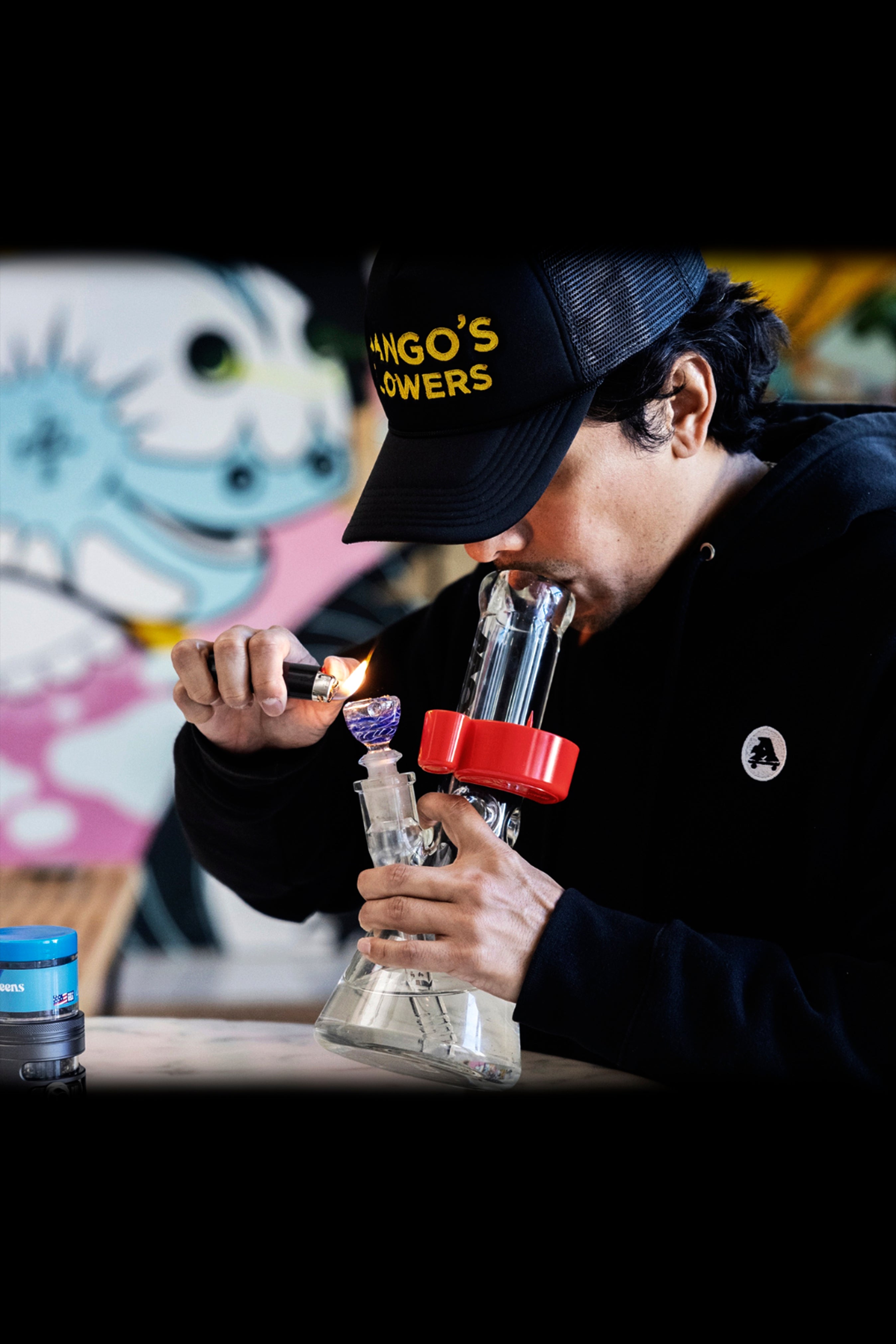 A person using a glass water pipe, illuminated by a lighter flame, while wearing a "Mango Flowers" branded hat.