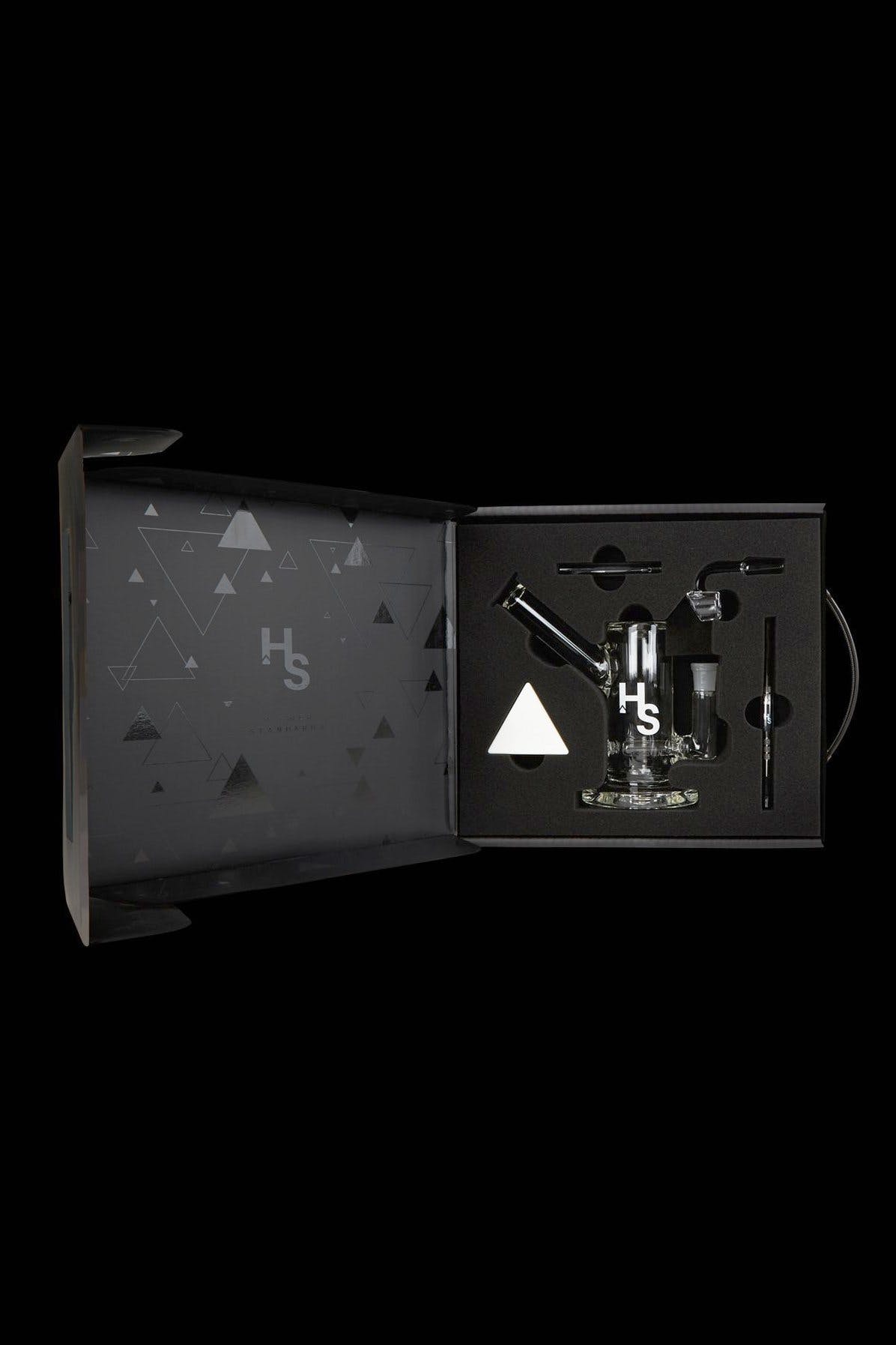 Open black gift box displaying cannabis smoking accessories, including a glass pipe, vaporizer, and tools, with a geometric pattern and "Hs" logo.