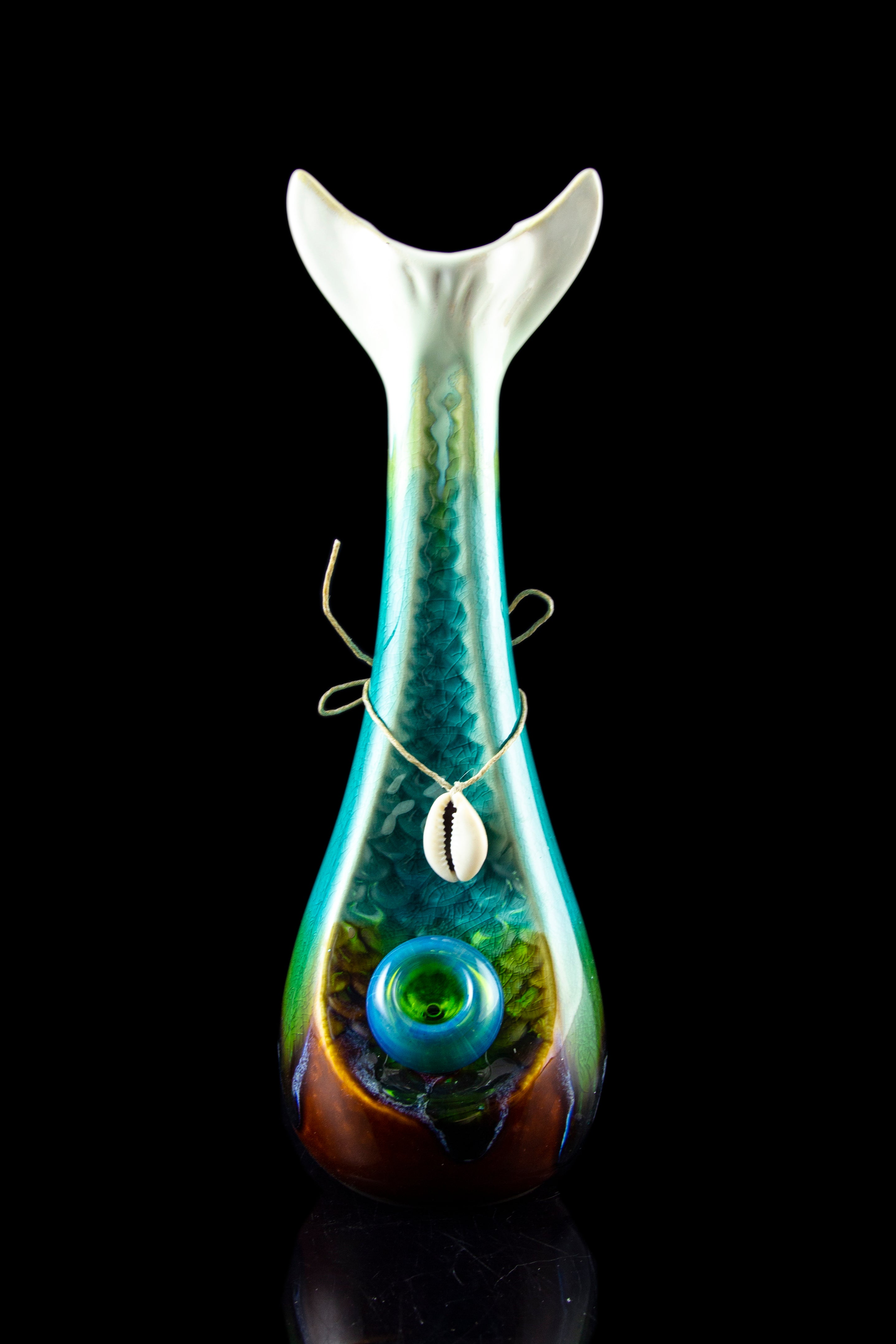 Hand-blown glass water pipe with teal and amber swirls, featuring a green sphere and adorned with a silver wire and cowrie shell pendant.