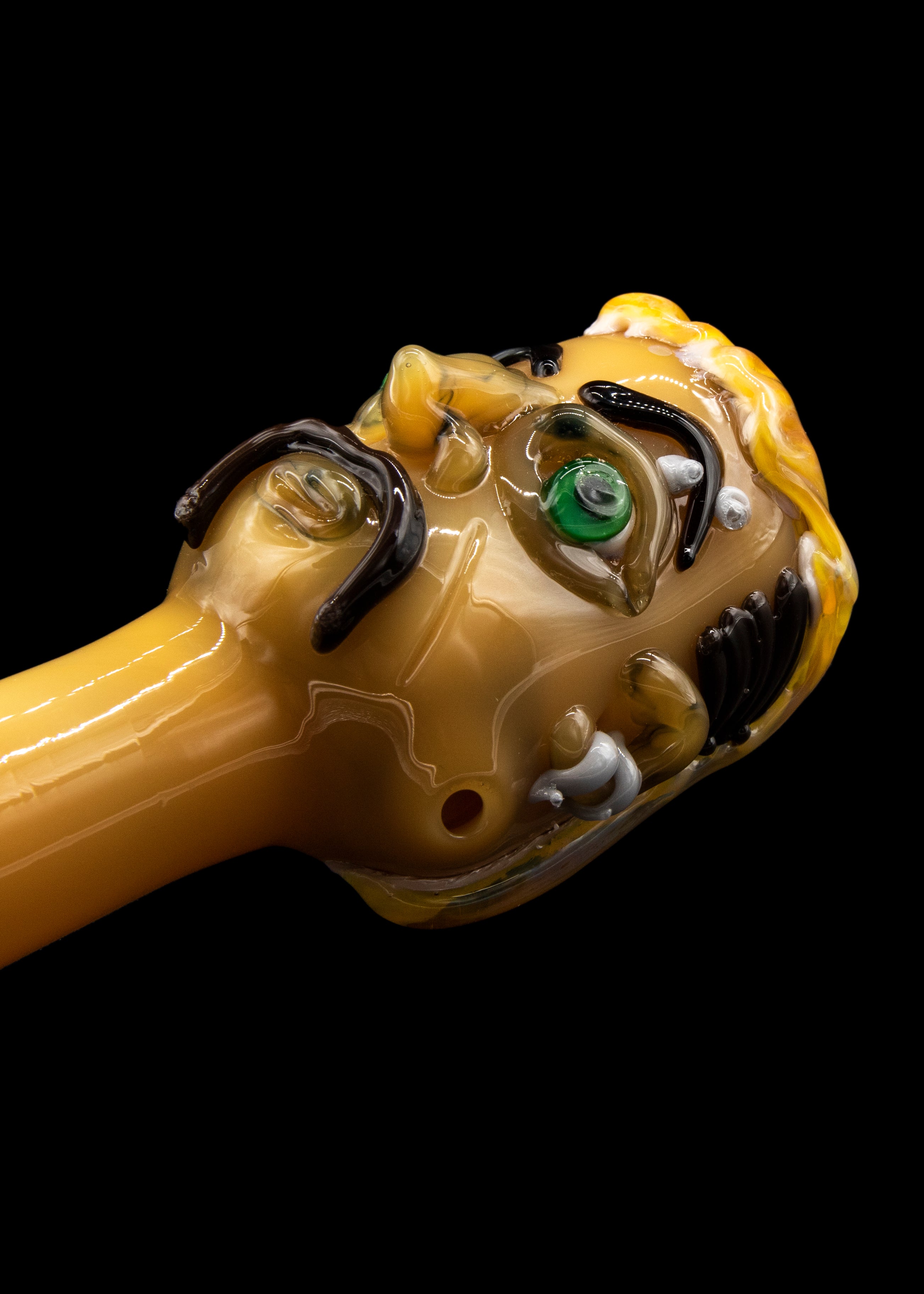 Hand-blown glass cannabis pipe sculpted as a grotesque face with a single green eye and swirling amber details.