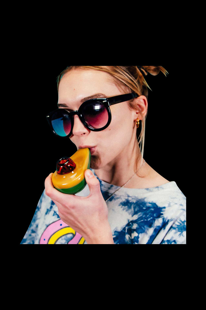 Close-up of a woman in sunglasses holding and using the Goody Avocado Hand Pipe, showing its yellow-green glass shape and red bowl.