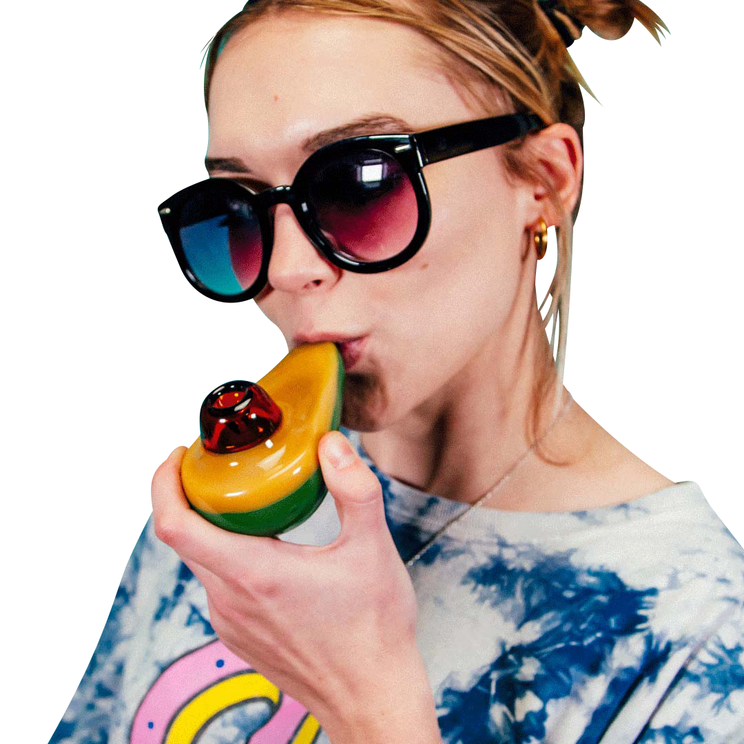 Close-up of a woman holding and bringing the Goody Avocado Hand Pipe to her lips against a teal backdrop, wearing dark sunglasses and a tie-dye shirt.
