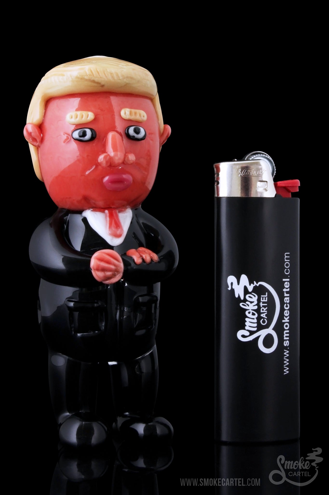 A sculpted Donald Trump figurine stands beside a black Smoke Cartel lighter, both against a dark background.