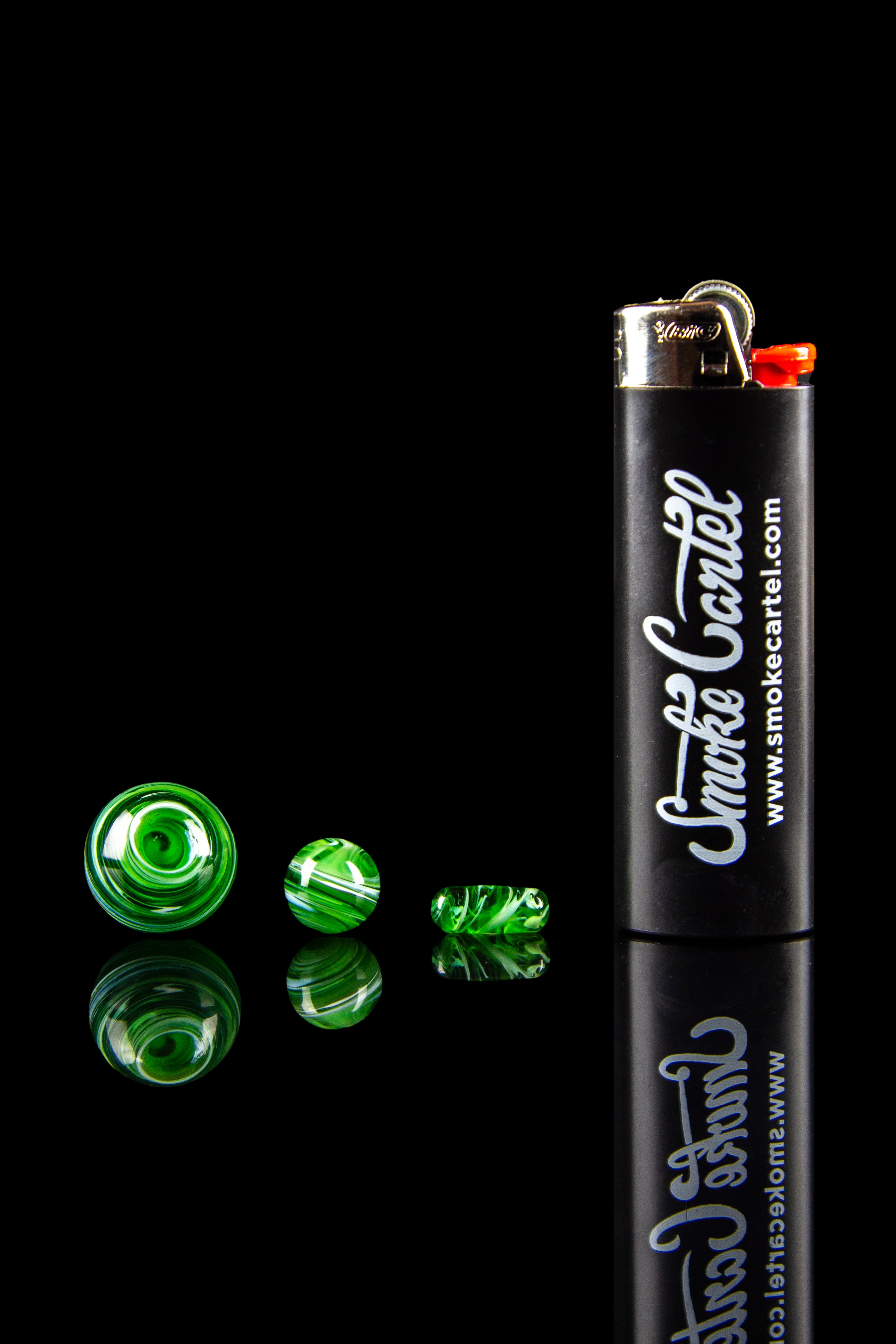Black lighter with "Smoke Cartel" branding and scattered green cannabis gems on a reflective surface.