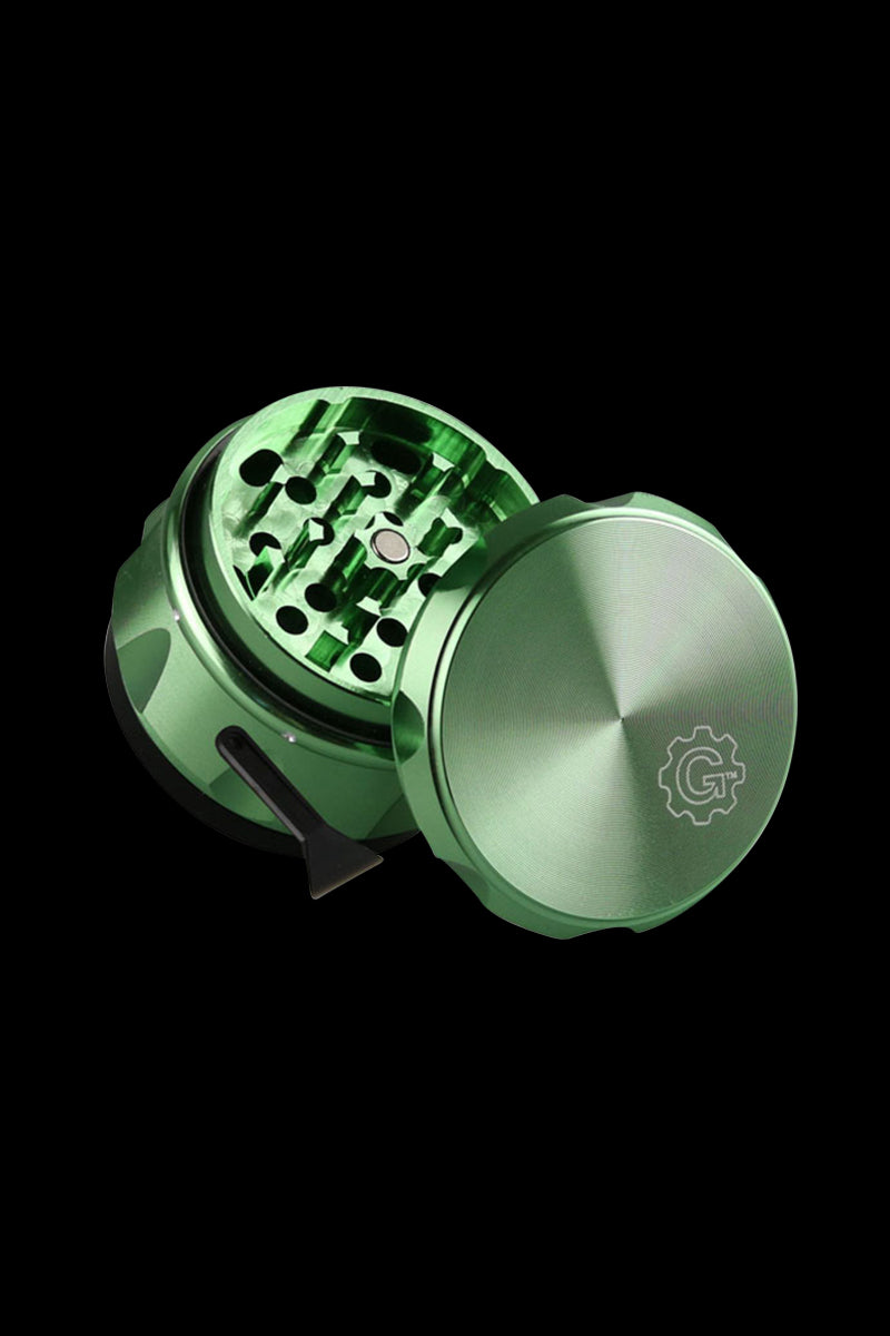 Angled close-up of the green Pulsar Carver 4-Piece Grinder showing its open, toothed grinding chamber, lid with logo, and small scraper on a black background