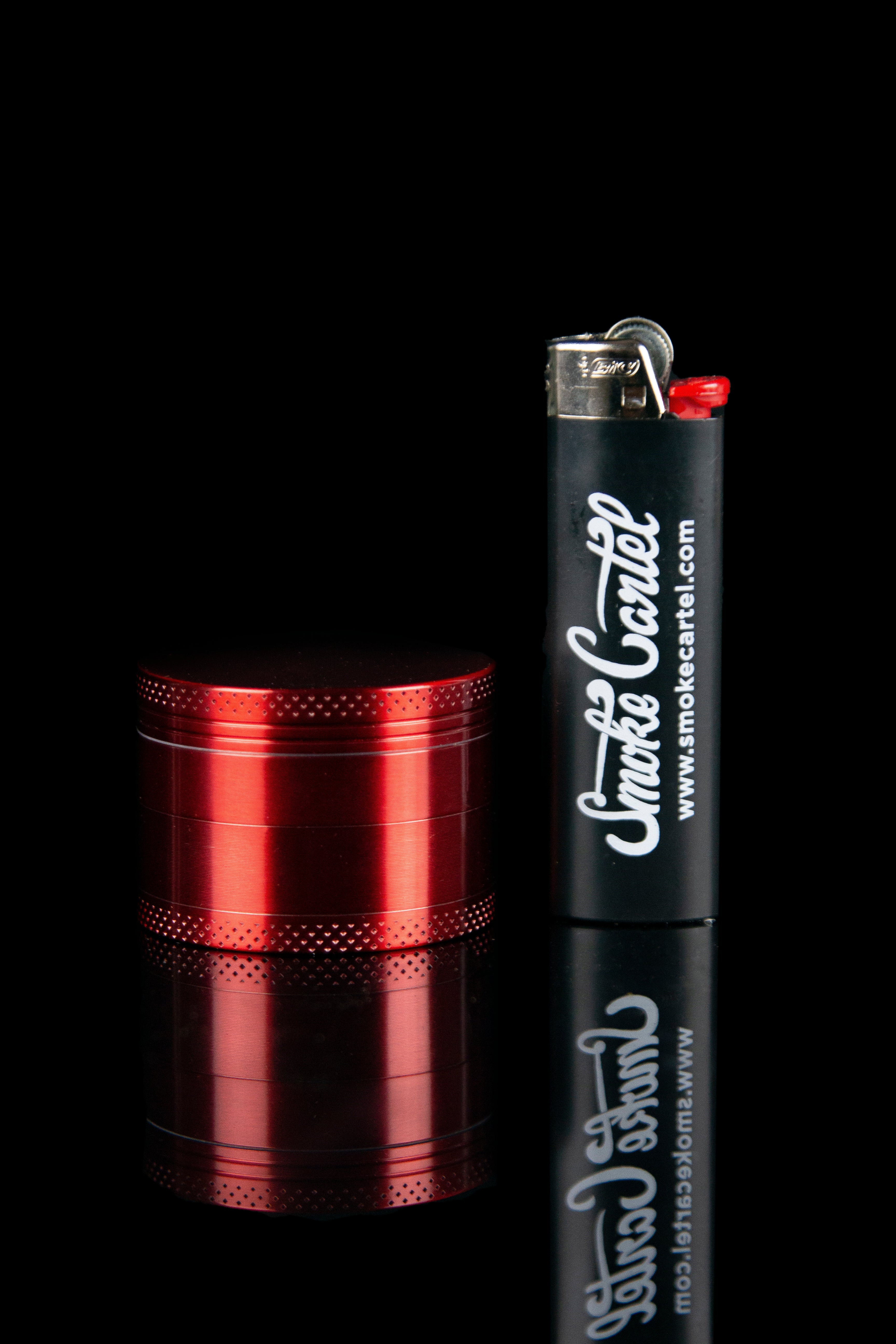 Red grinder and lighter with “Smoke Cartel” branding reflected on a black surface.