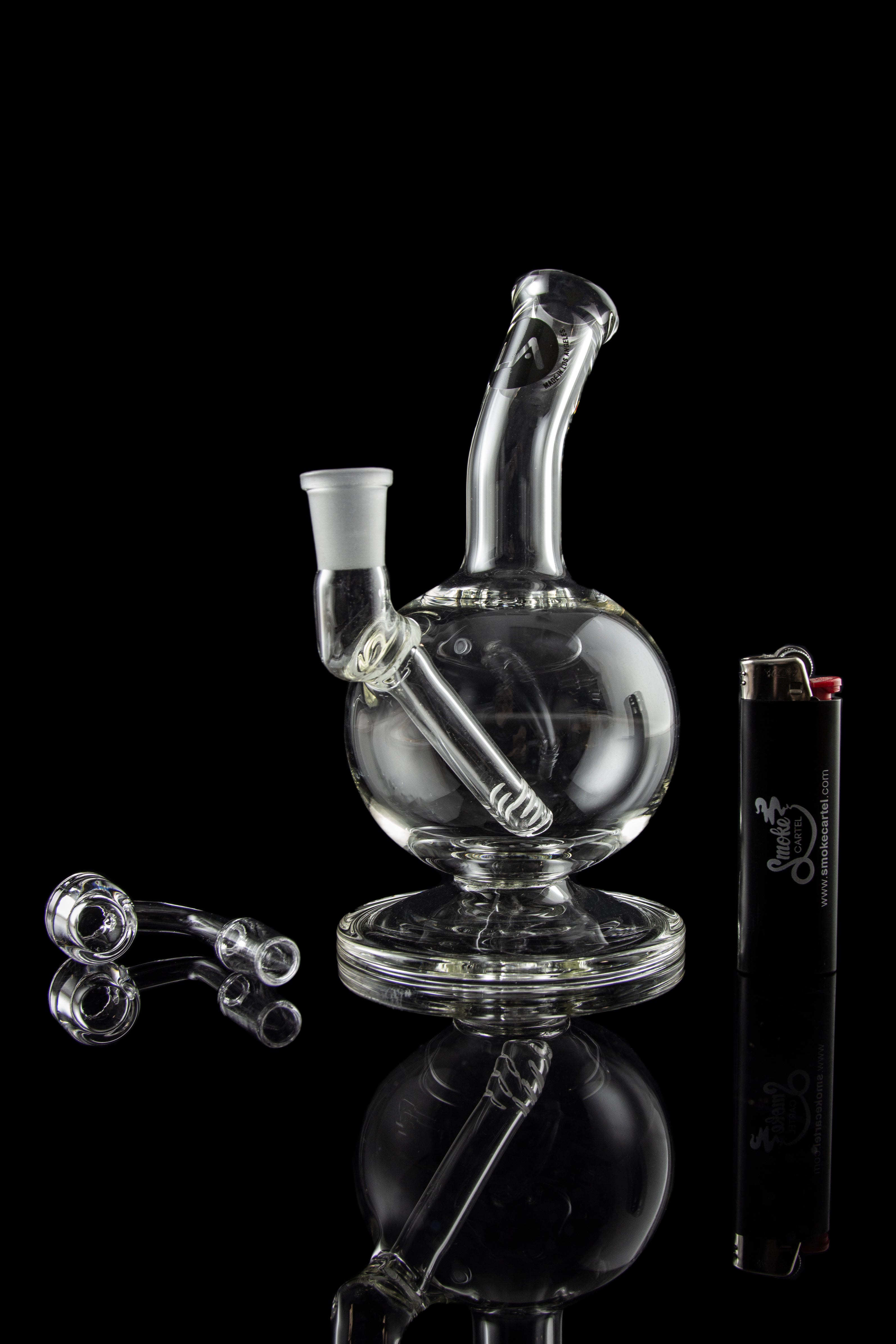 Clear glass water pipe (bubbler) with accompanying glass pieces and lighter, studio shot against a black background.