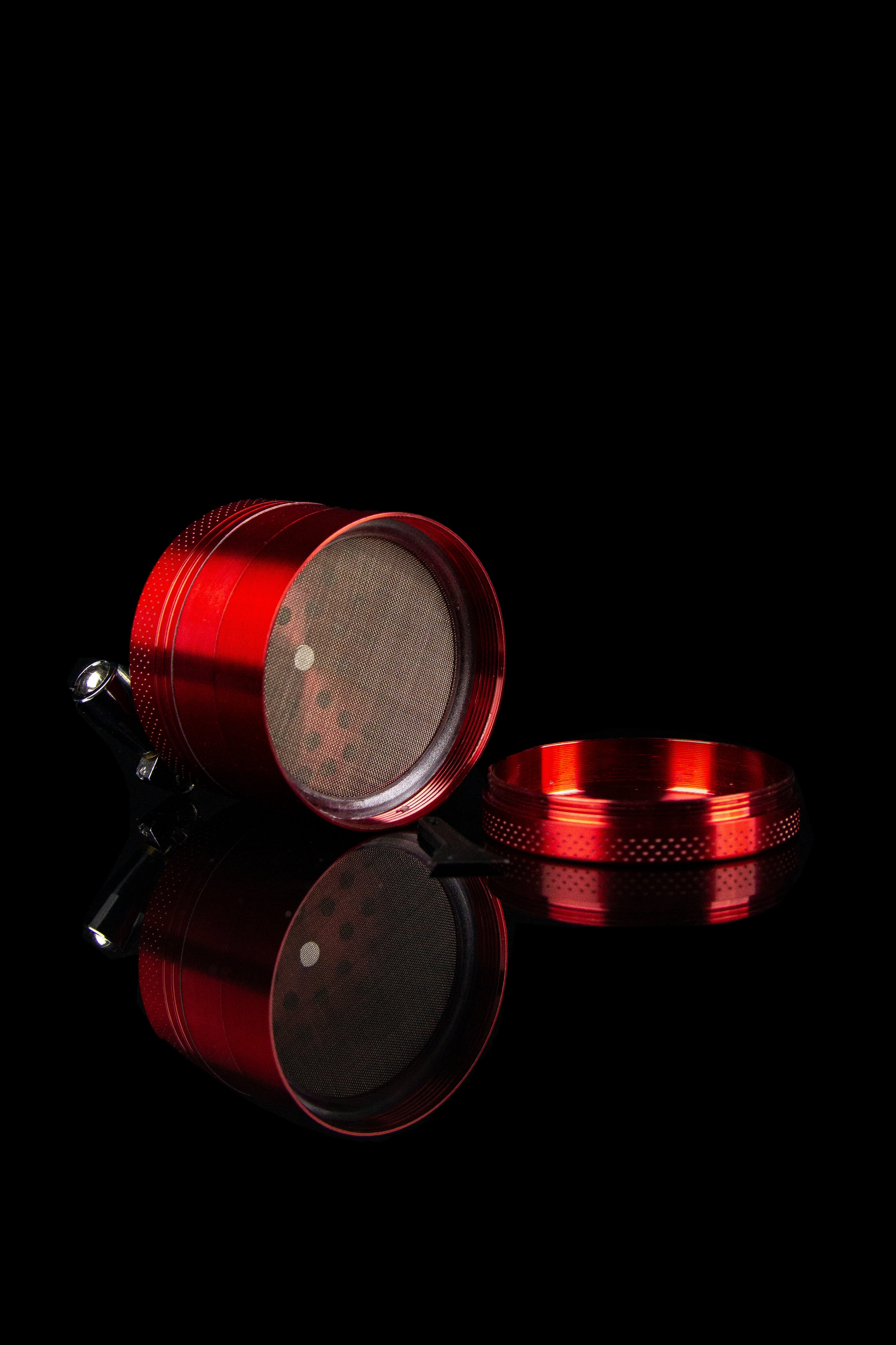 Red cannabis grinder with open components and reflective surface, isolated on black.