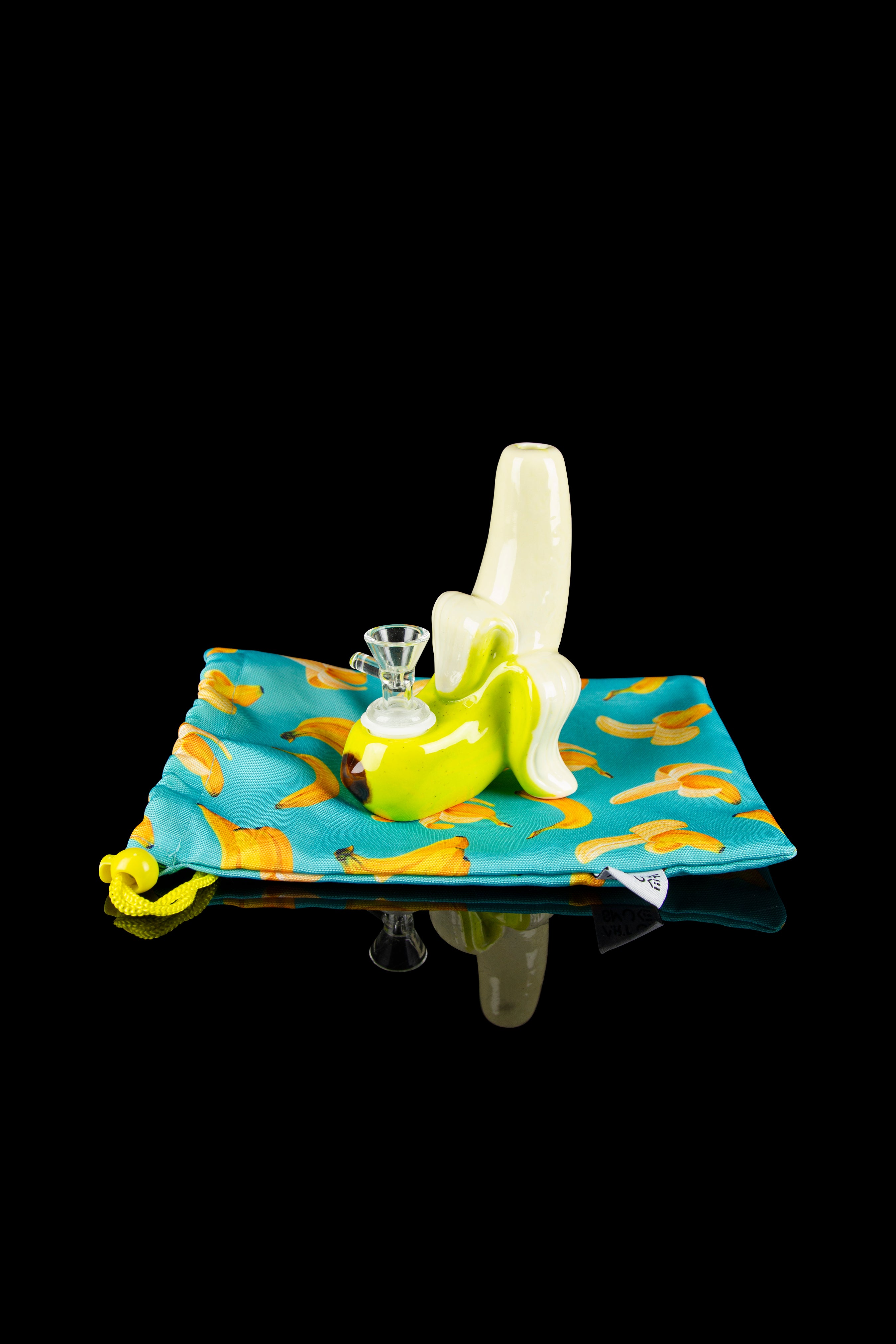 A sculpted glass banana-shaped water pipe rests on a patterned pouch, with a second banana piece lying beside it.