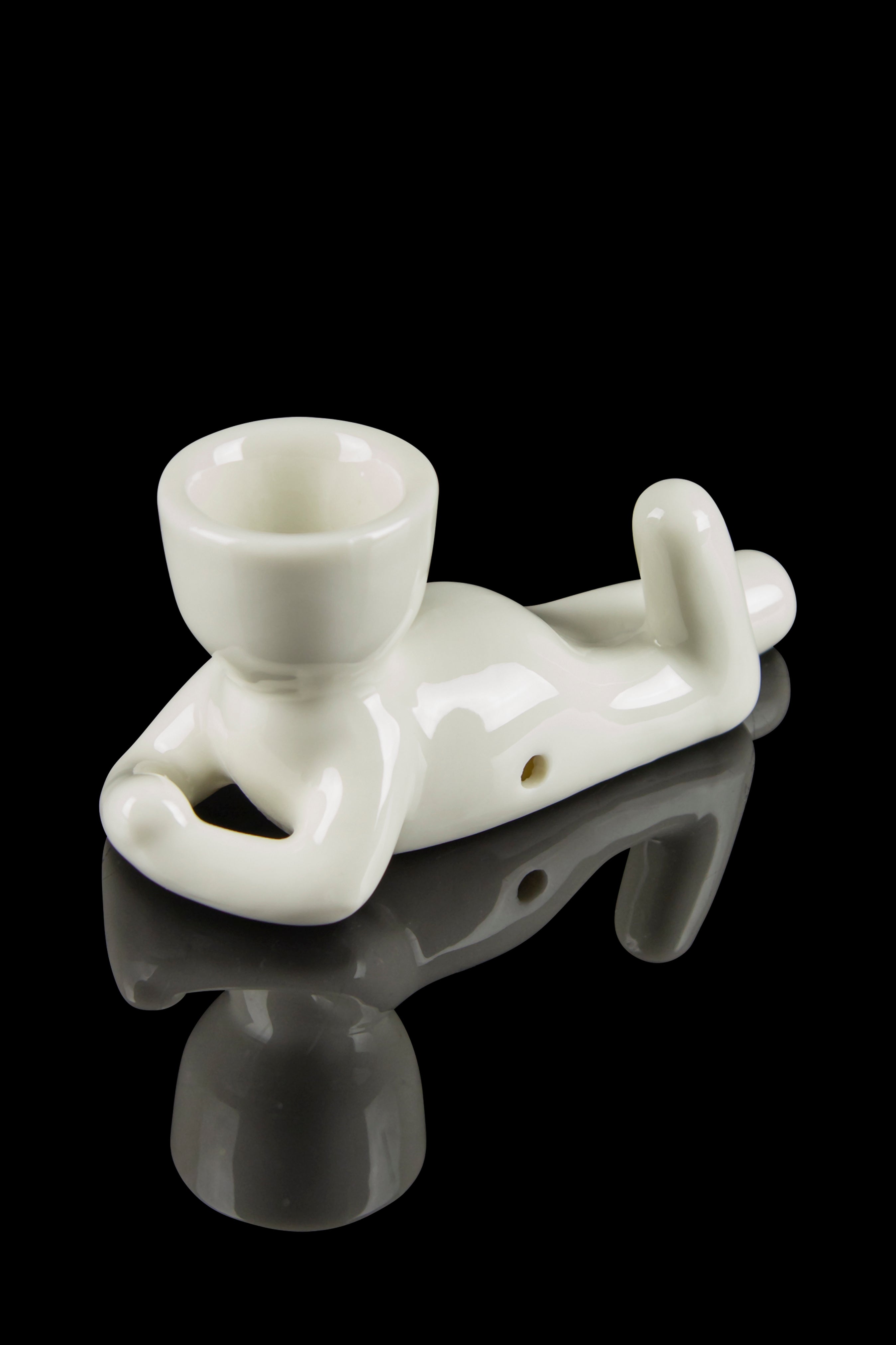 White ceramic chillum pipe shaped like a lounging figure, reflected on a black surface.