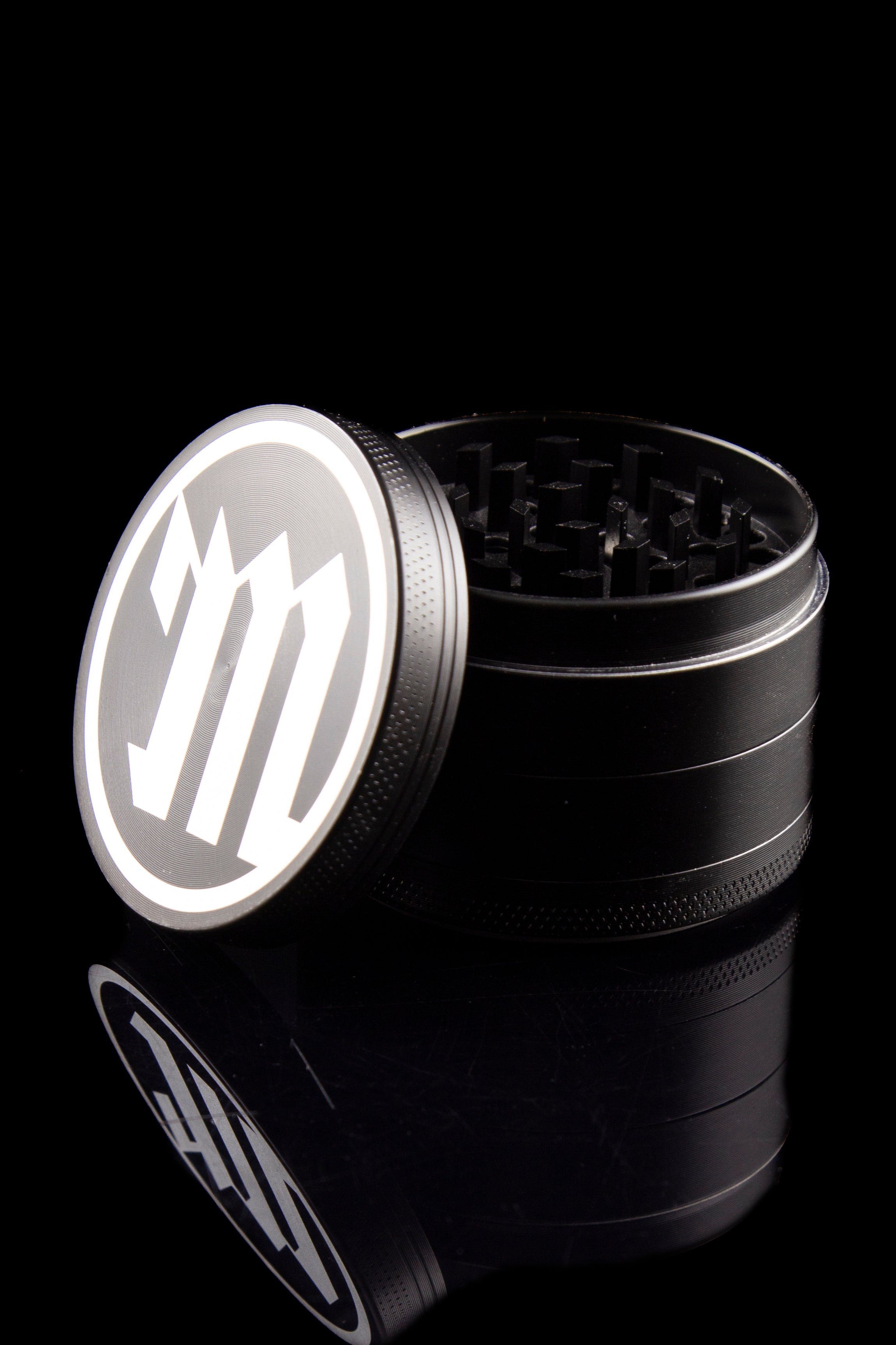 Black metal cannabis grinder with a branded lid, partially disassembled, against a dark reflective surface.