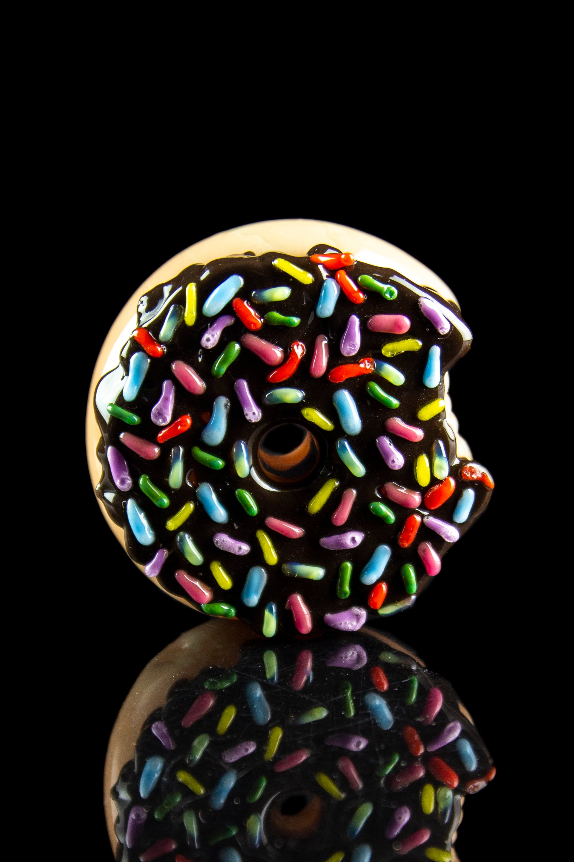 Glass pipe shaped like a chocolate donut with colorful sprinkles, reflected on a black surface.