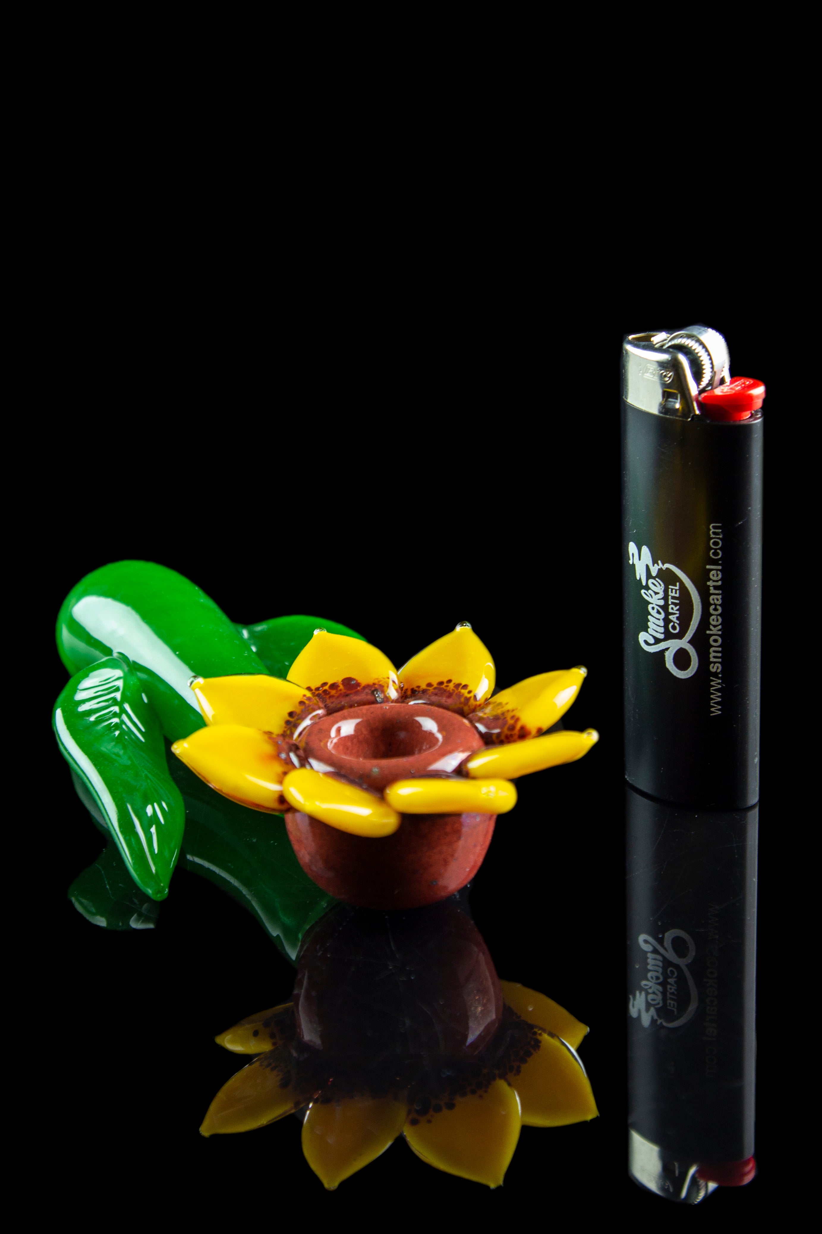A vibrant green and yellow glass flower pipe rests beside a black lighter branded with "Smoke Cartel" on a reflective black surface.