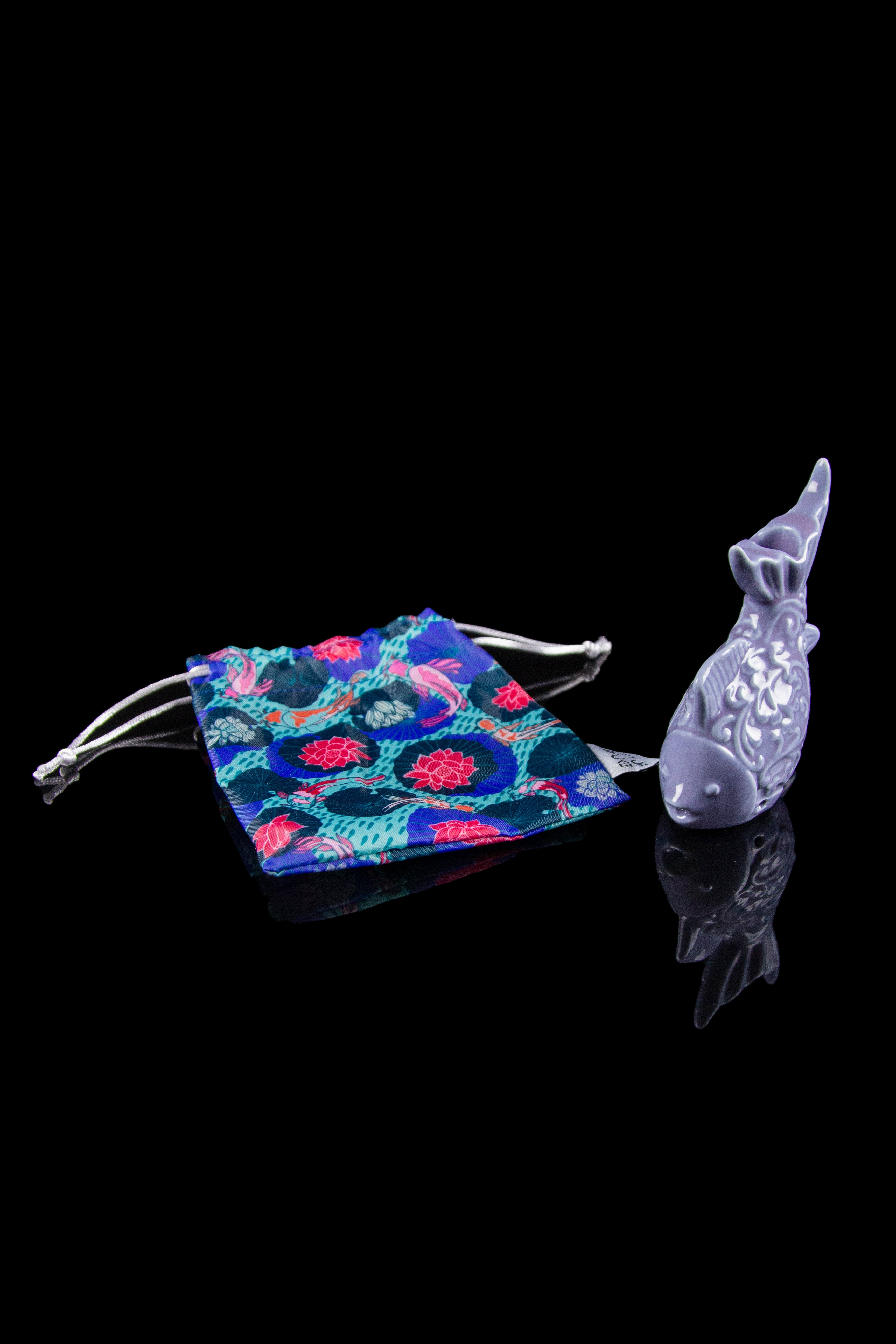 Ceramic fish-shaped smoking piece next to a patterned velvet pouch on a black background.