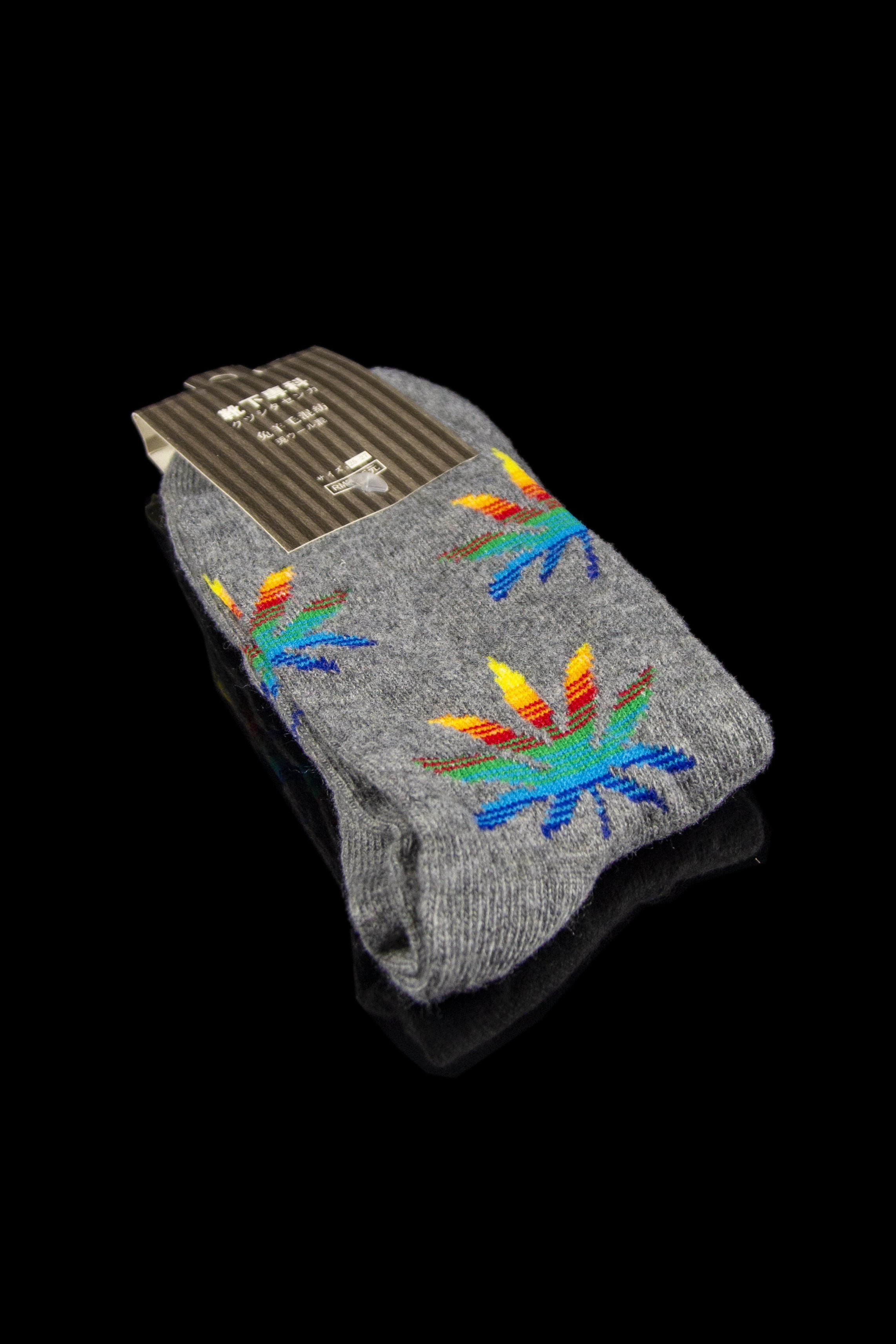 Folded gray socks with colorful cannabis leaf patterns, still attached to a cardboard tag, against a black background.