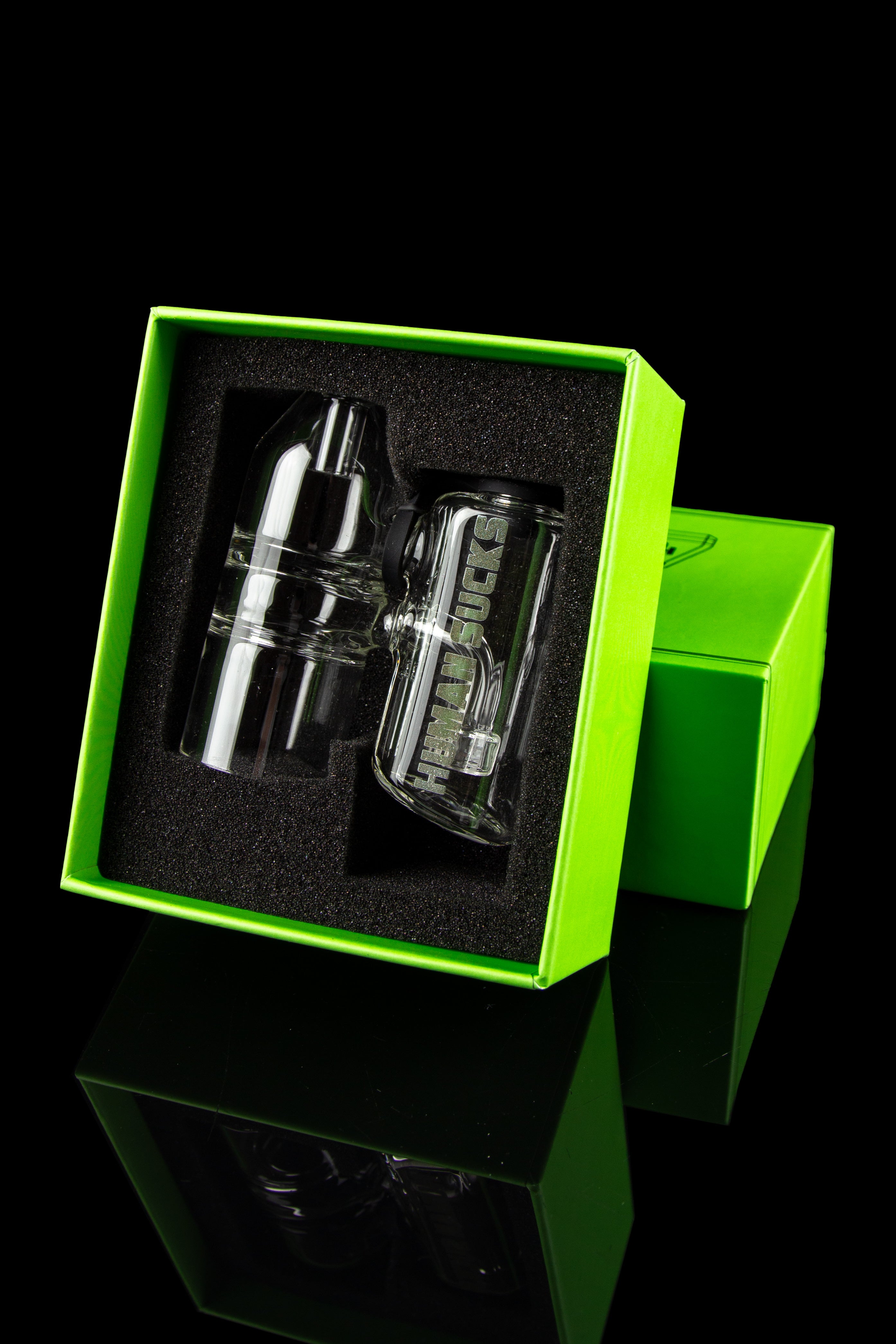 A clear glass cannabis water pipe with "Human sucks" etched on the side, displayed in an open bright green gift box with a black foam insert.