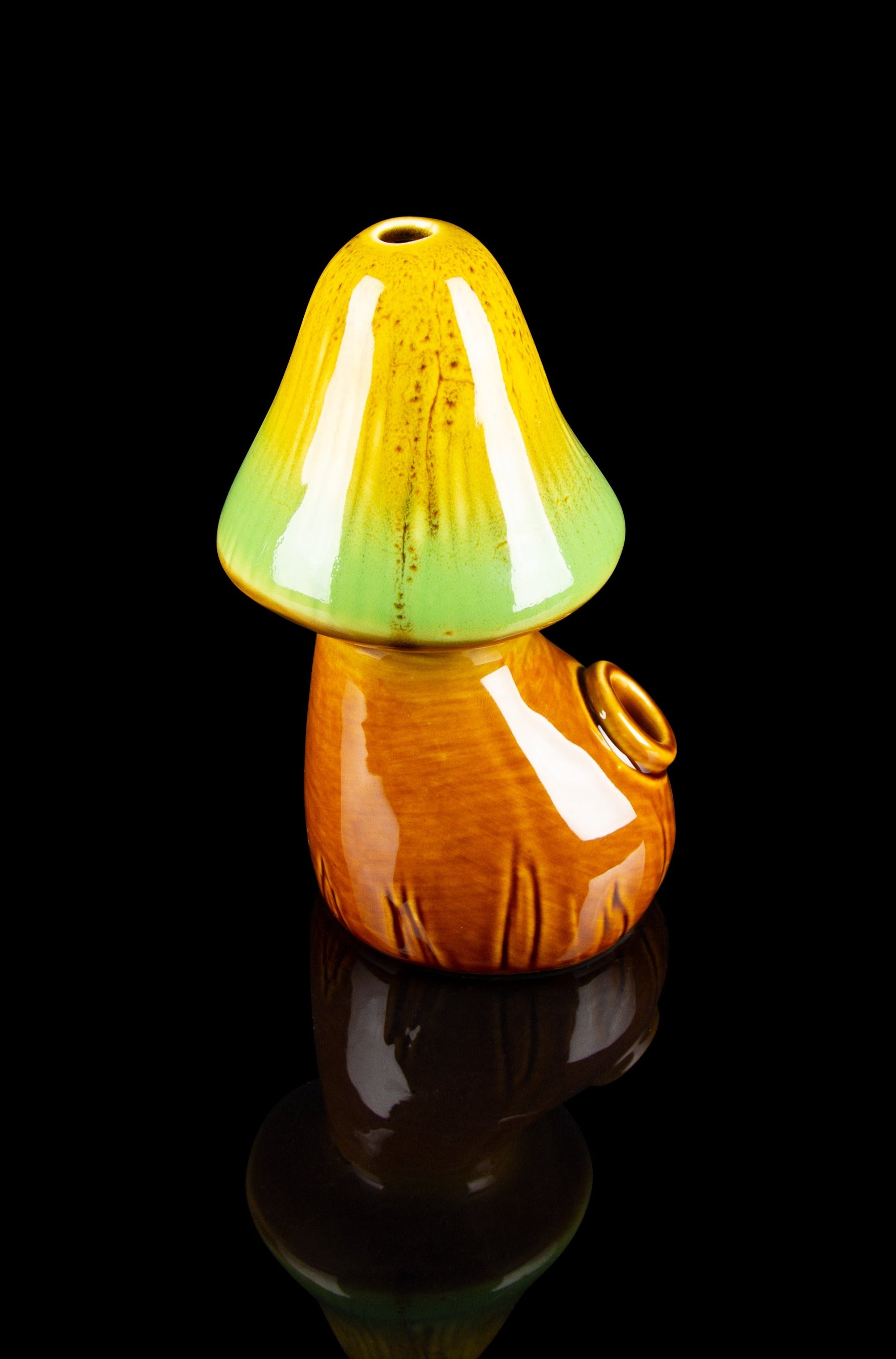 Ceramic mushroom-shaped cannabis pipe with a glossy, orange and green glaze, reflecting on a dark surface.
