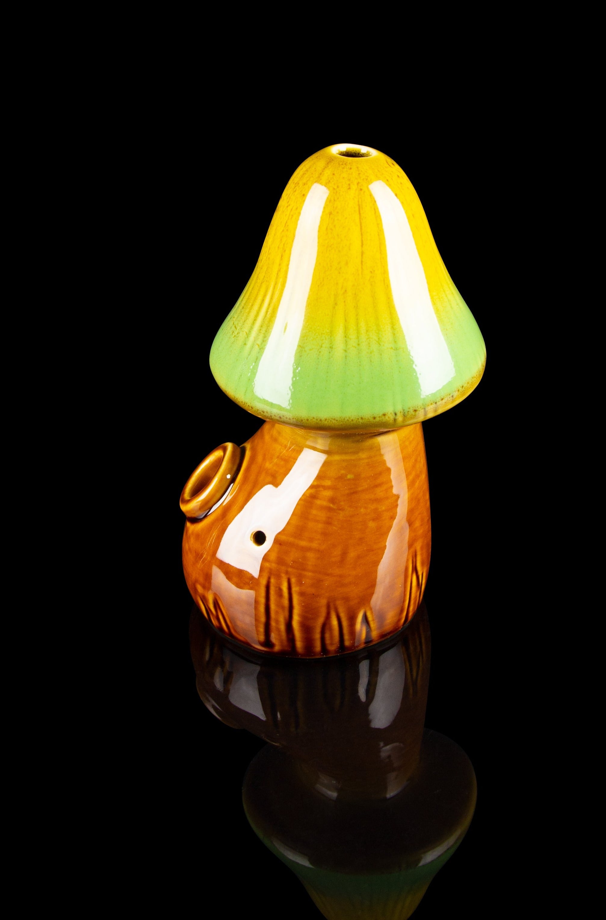 Ceramic mushroom-shaped cannabis pipe with orange and green glaze, displayed on a reflective surface against a black background.