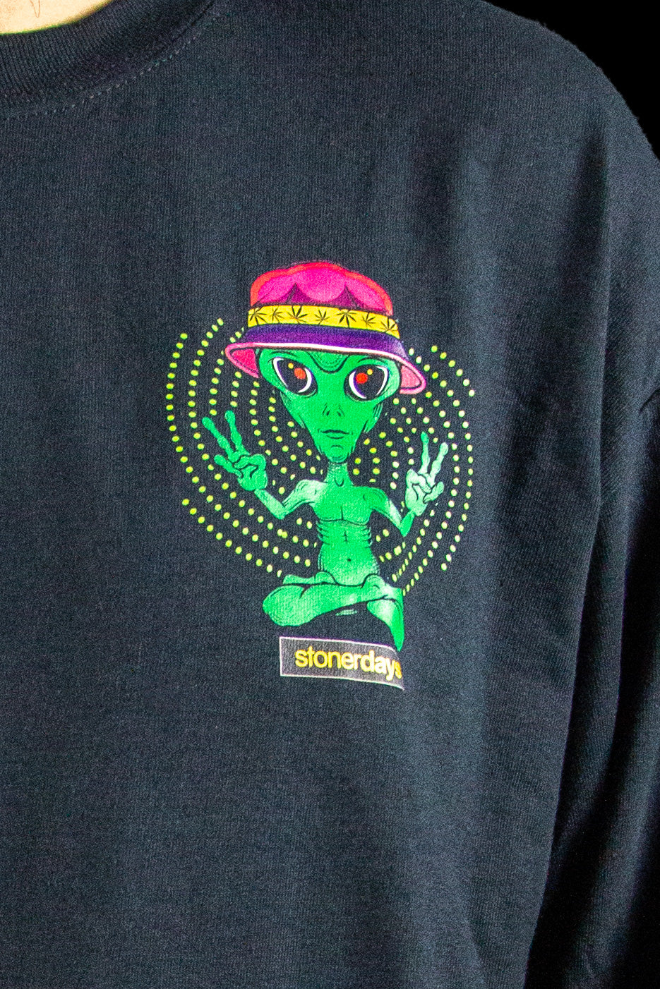 Close-up of a black sweatshirt featuring a vibrant, psychedelic print of an alien meditating in the lotus position with the text “stonerday” below.