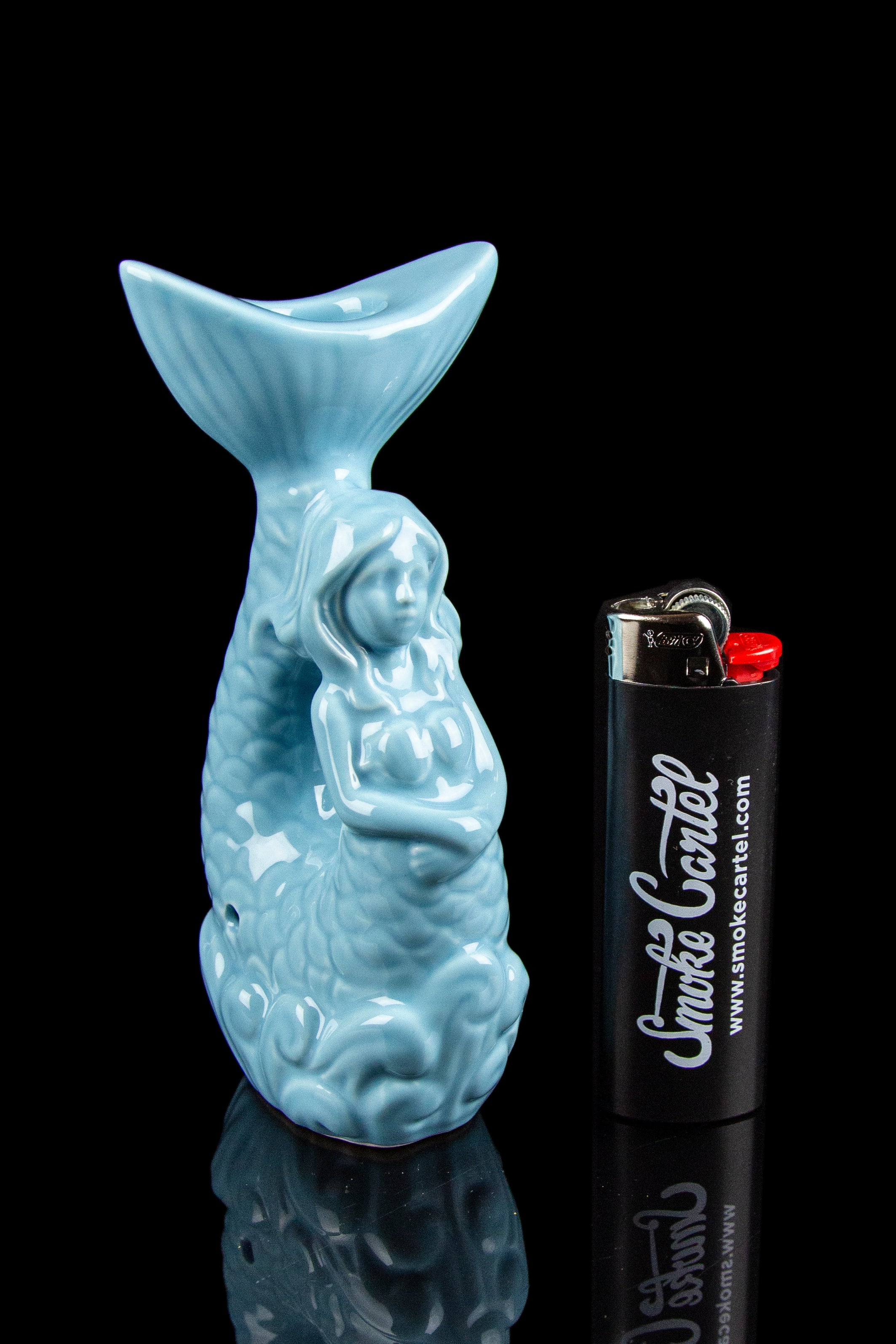 Blue ceramic water pipe shaped like a mythical creature, next to a white lighter with "Smoke Cartel" branding, reflected on a glossy black surface.