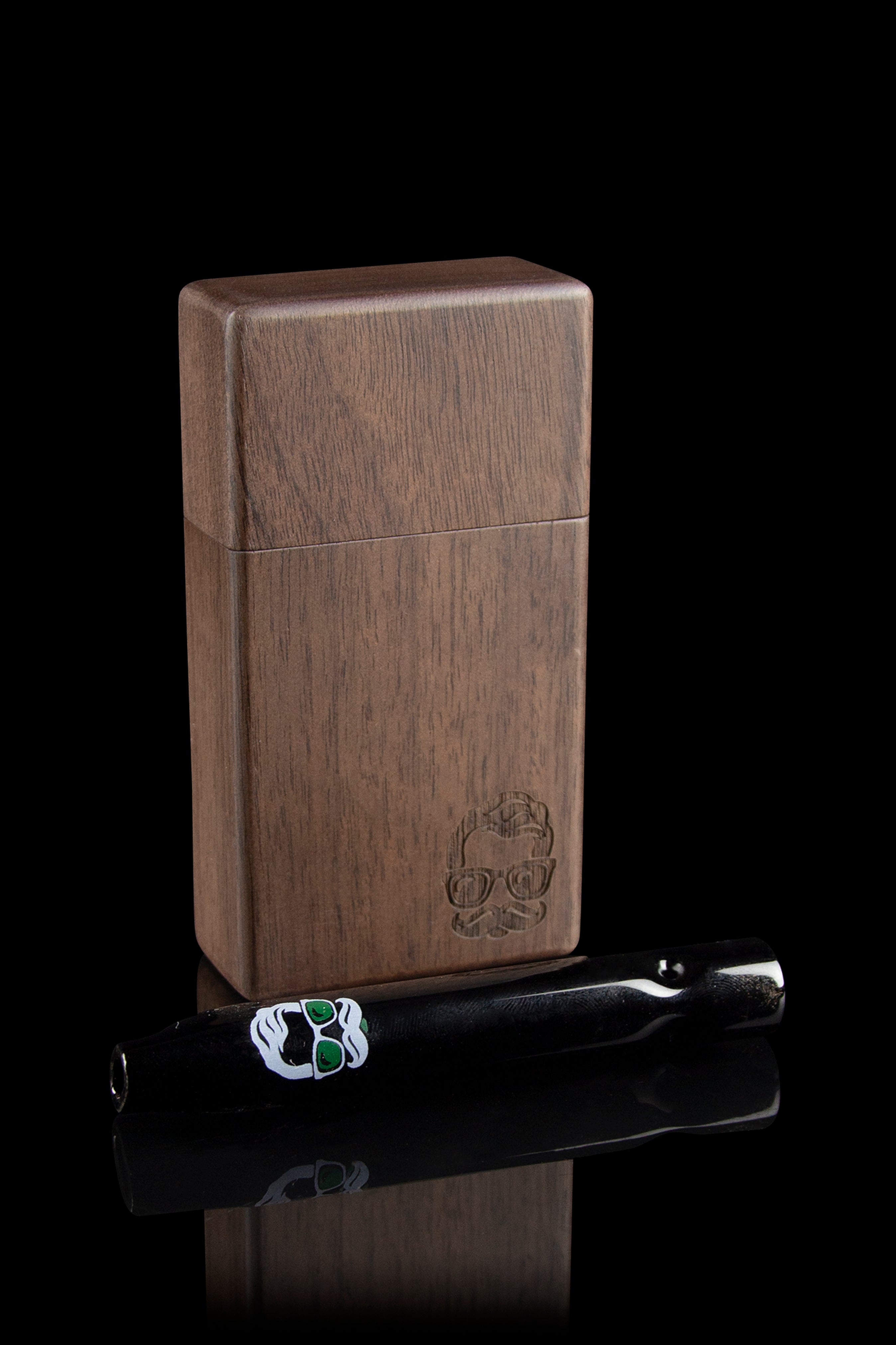 Wooden cannabis smoking box with a skull design and glass pipe resting on a reflective surface.