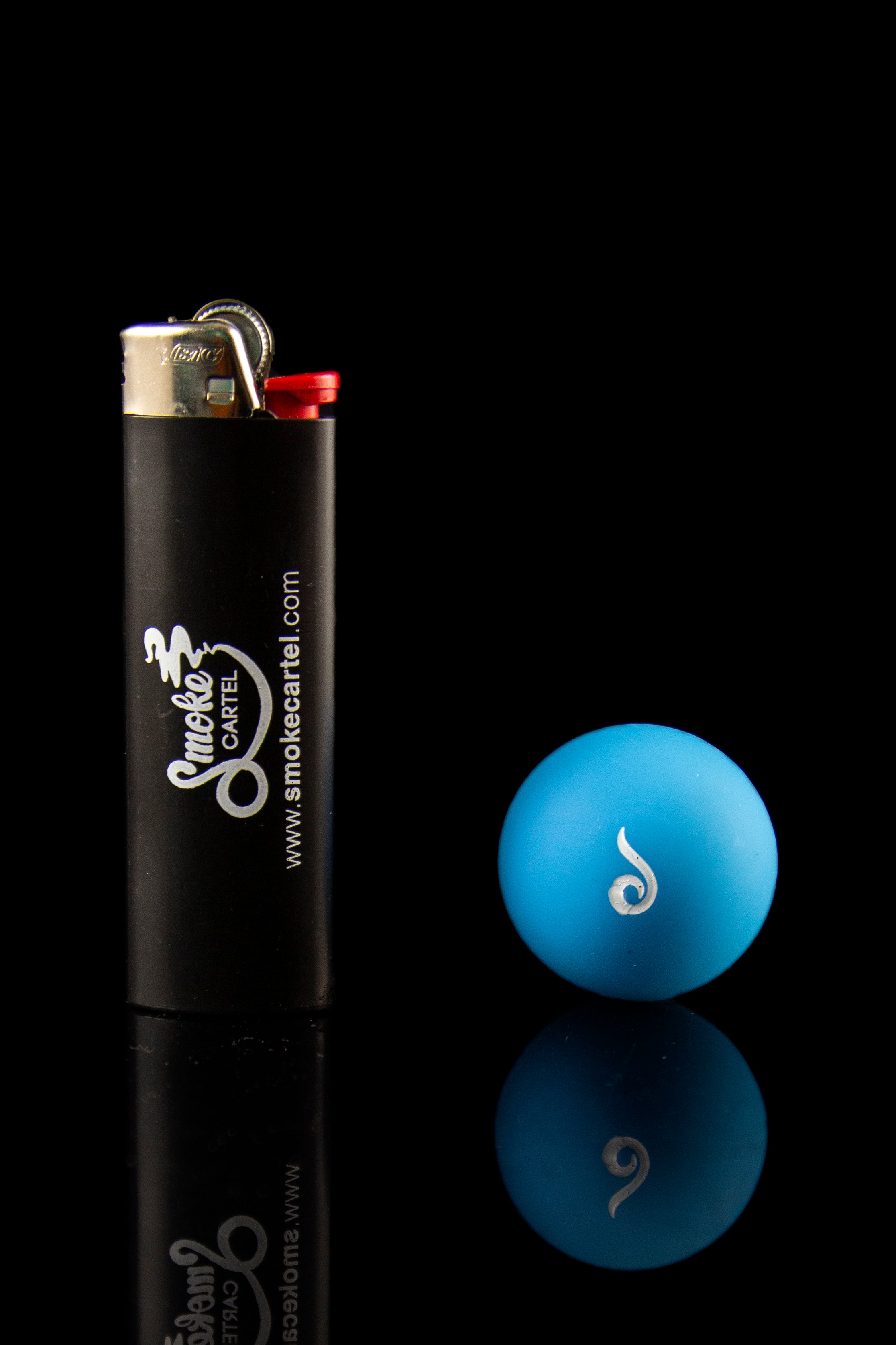 Black lighter with "Smoke Cartel" branding next to a blue glass marble with white swirl design, both reflected on a dark surface.