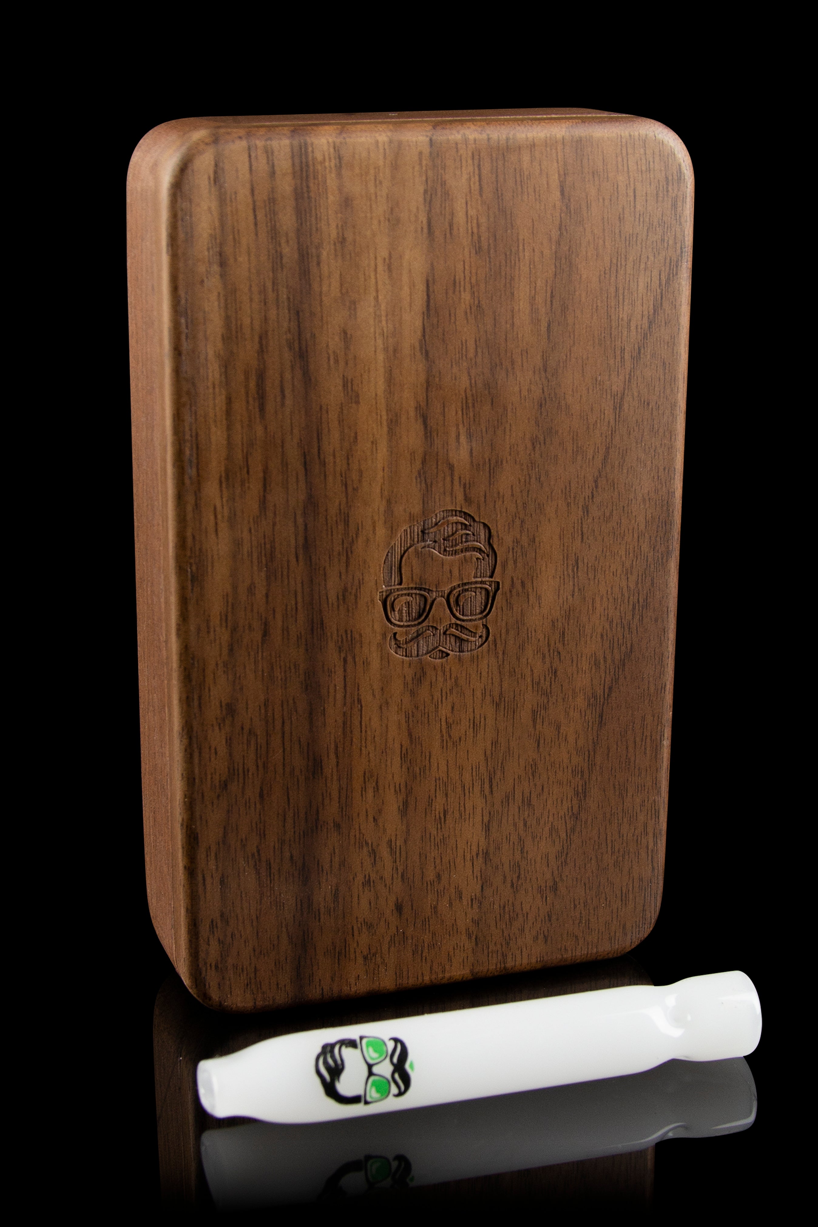 Wooden cannabis smoking case with engraved skull design, alongside a white one-hitter pipe on a reflective surface.