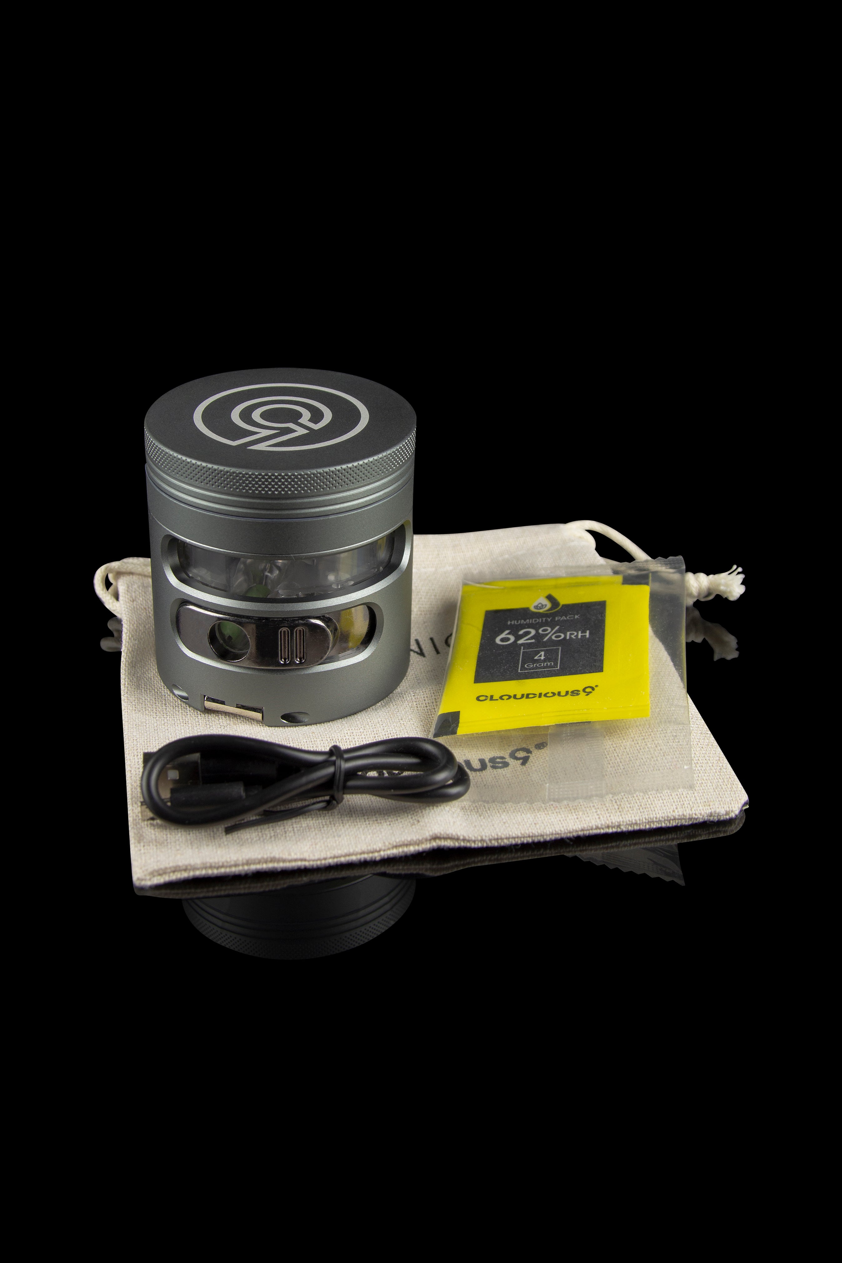 Silver cannabis grinder, humidity pack, and USB charging cable on a tan linen pouch against a black background.