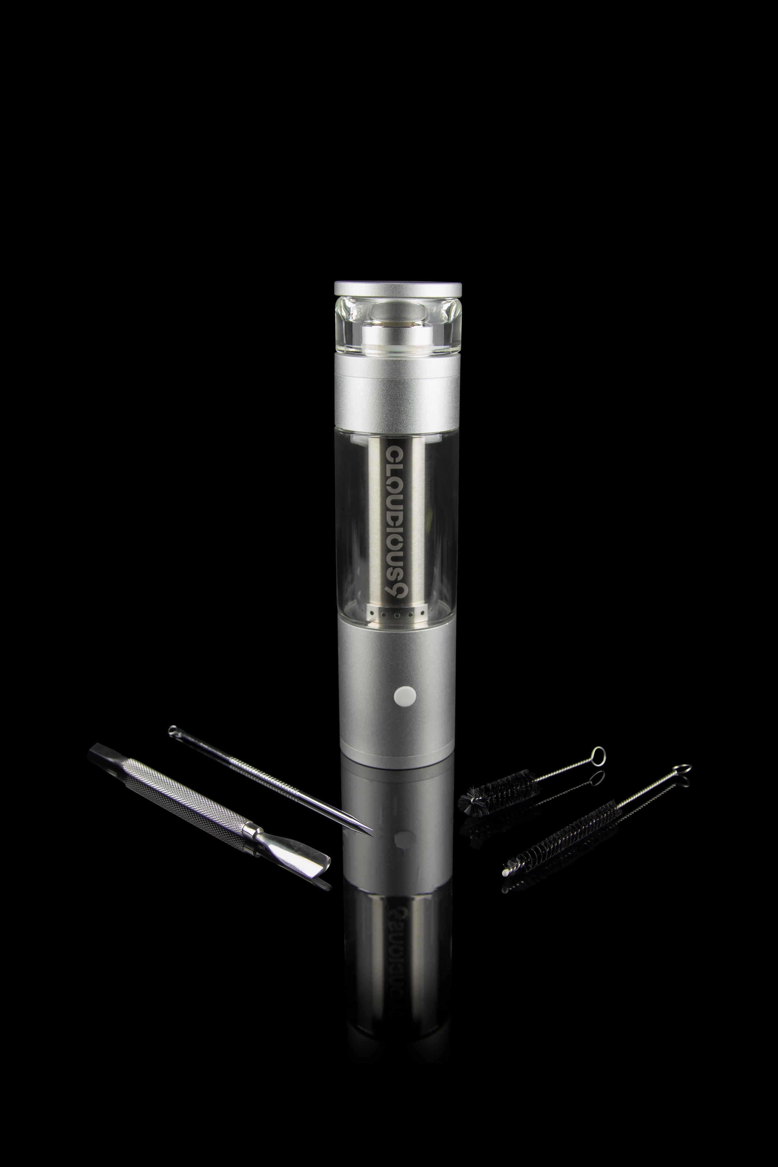 Silver cannabis vaporizer with glass chamber and cleaning tools on a reflective surface.