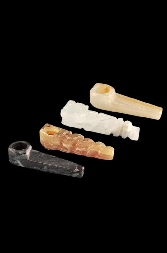Angled overhead view of four Onyx Stone Smoking Pipe pieces—black, amber, white, and beige—arranged diagonally on a black background.