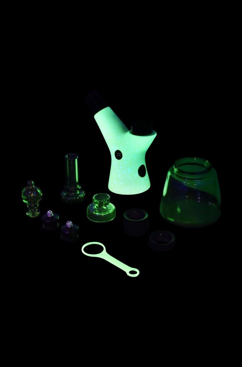 Glowing green glass cannabis water pipe and accessories on a black background.