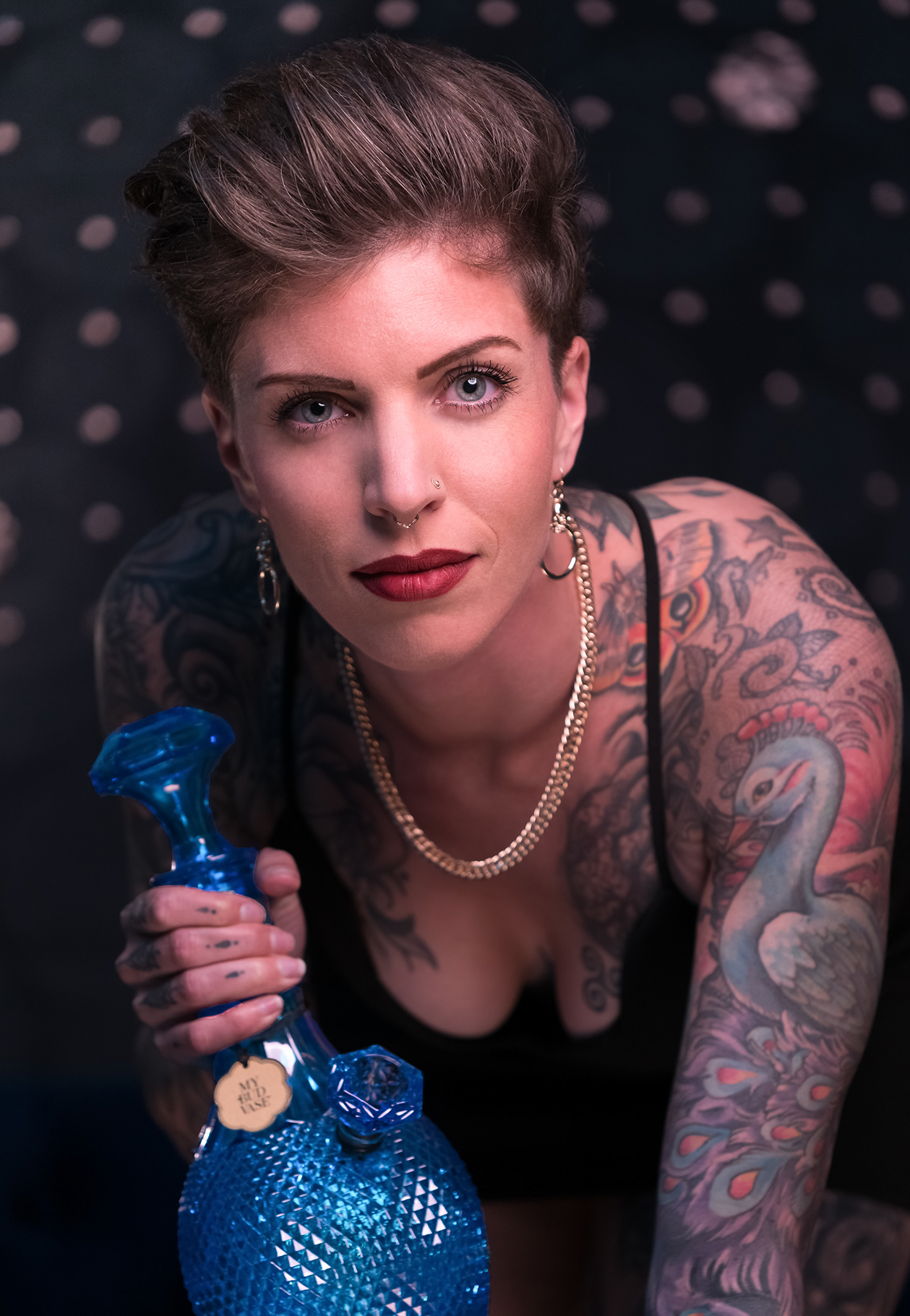 A woman with short hair and intricate tattoos gazes directly at the viewer, holding a blue glass water pipe.