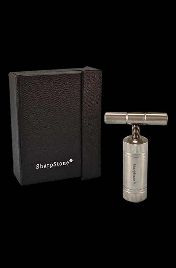 Studio-lit product shot of the Sharpstone T-Style Press with Case showing the stainless T-handle press standing upright beside its textured black SharpStone-branded box on a black background