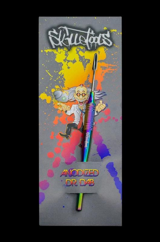 Vertical close-up of a rainbow anodized Skilletools Anodized Series Dab Tools "Dr. Dab" tool laid on colorful splatter packaging featuring a cartoon scientist.